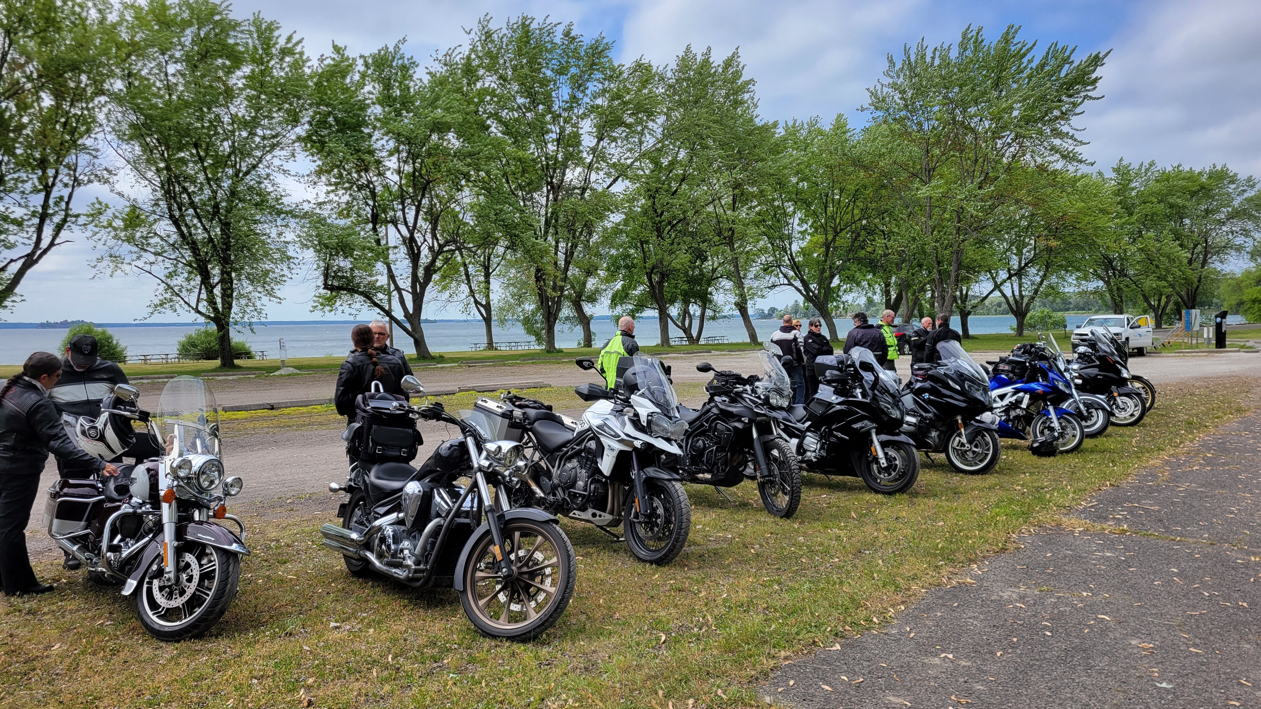 a row of motorcycles and riders parked in the grass at a rest stop along Ontario's Highway 2, by a treed lakefront on a summer day.