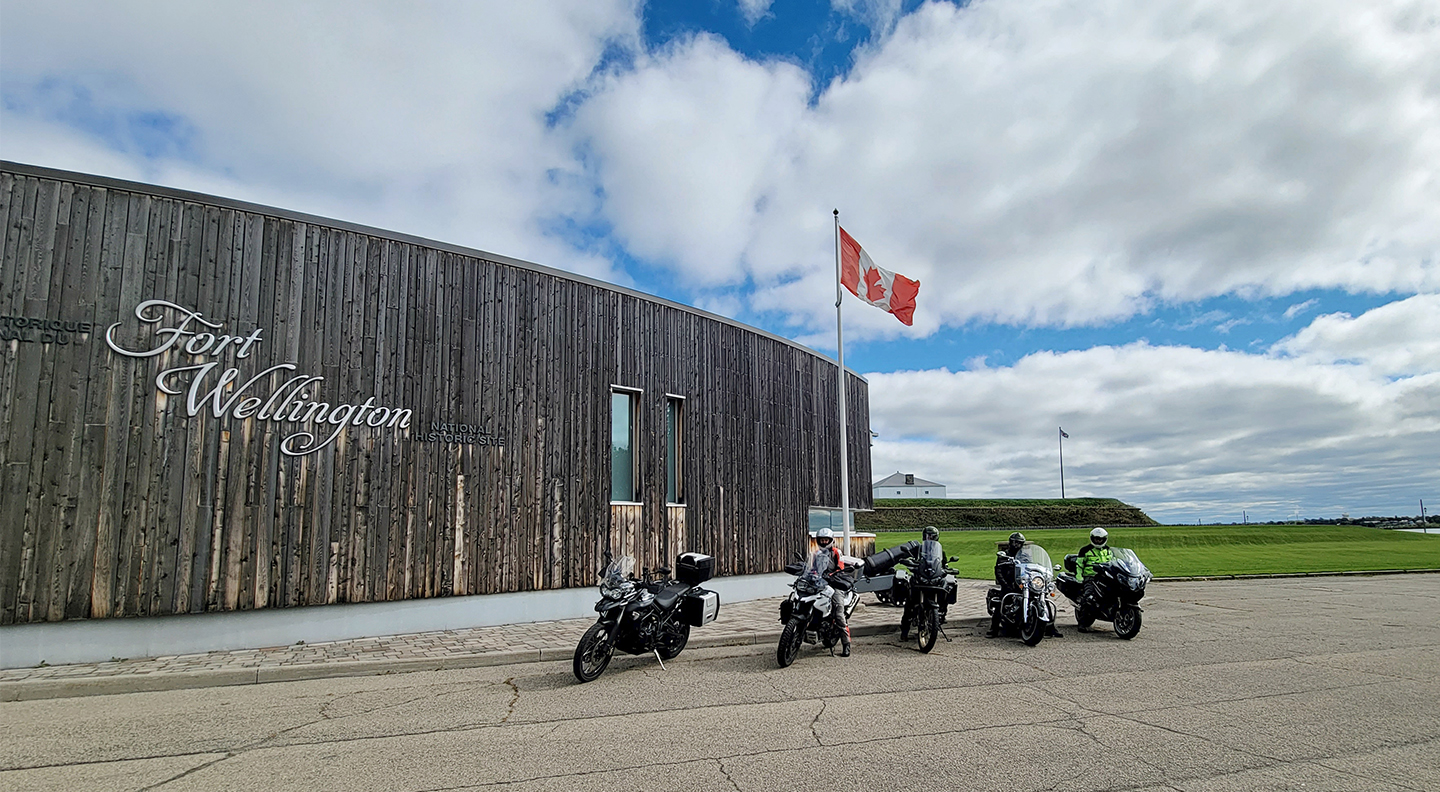 5 motorcycles parked in line in front of Fort Wellington, a fortress protected by a circle of tall fence of wooden poles shielding it from view. A Canada flag flies on a pole next to it.  