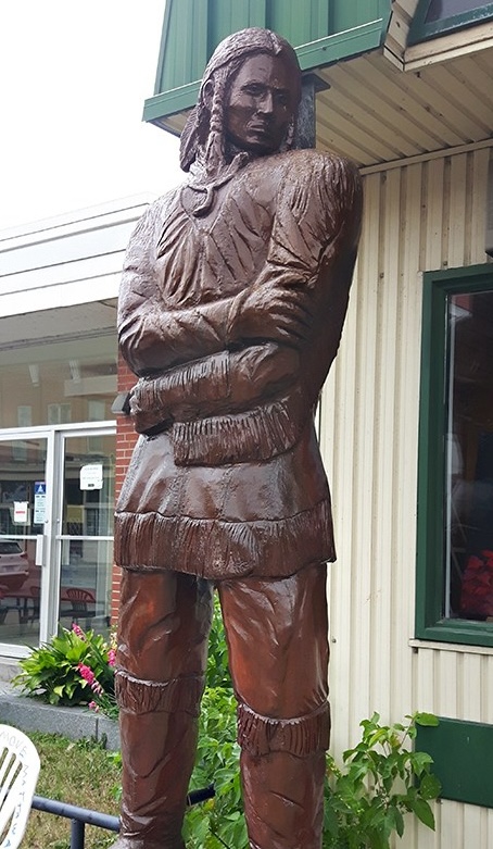 A tall carved wood statue of an indigenous man in Mattawa, standing outside a store.