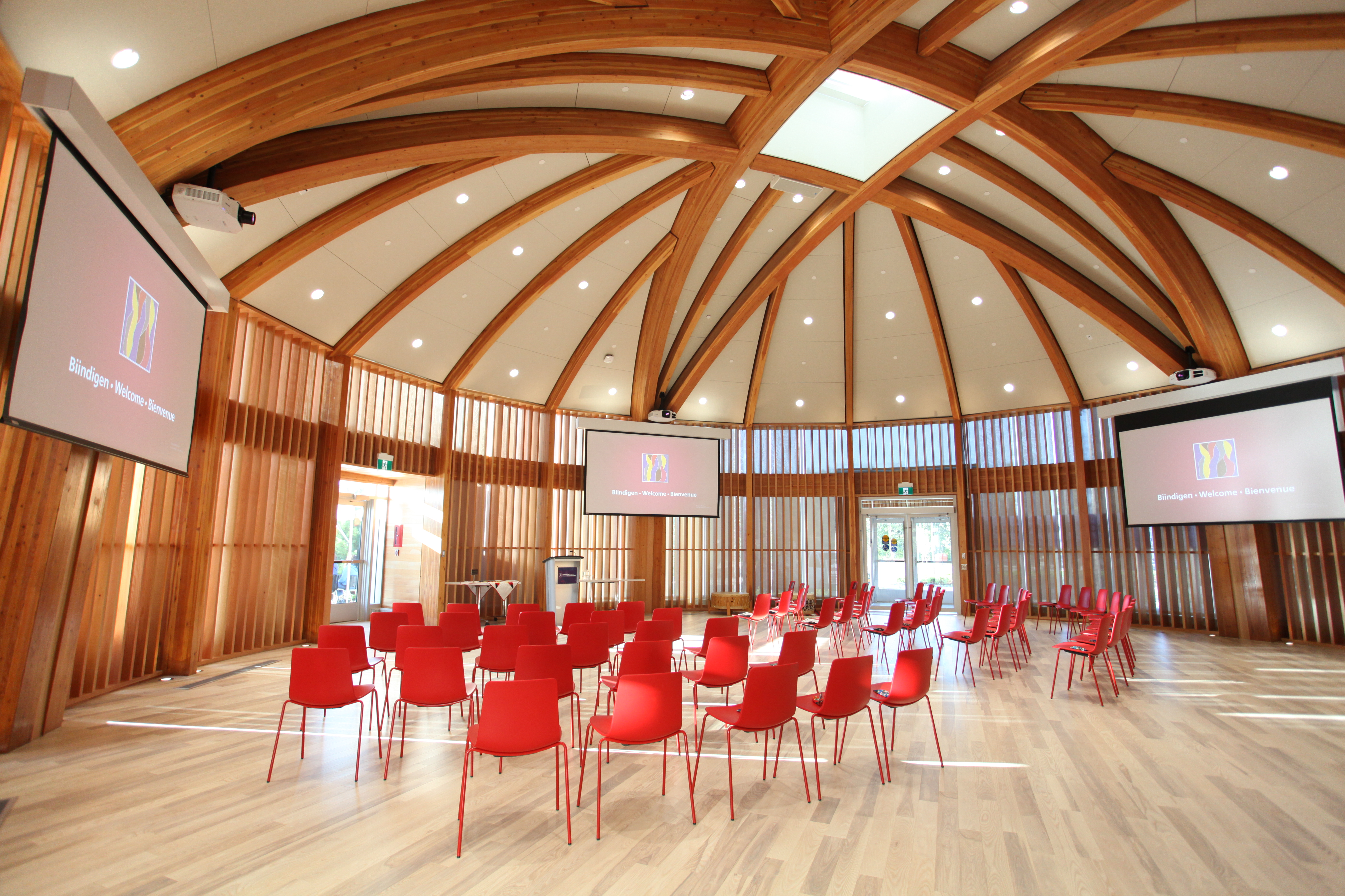 The Indigenous Sharing and Learning Centre; a large, well-lit, circular room with a vaulted ceiling, several display screens on the walls and two rows of red chairs in the centre.