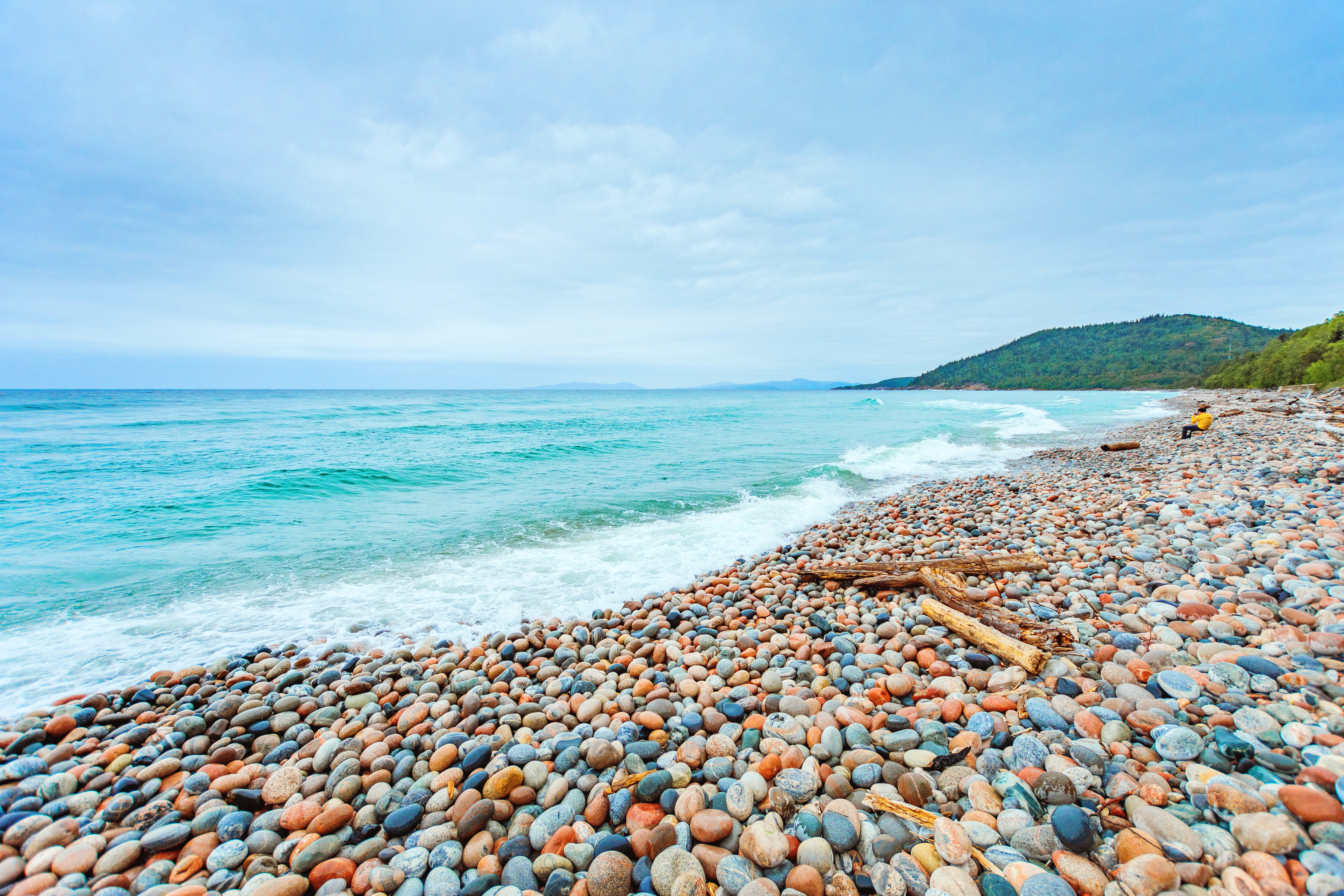 The waving turquoise water of Lake Superior meets a beach made of multicoloured round stones under a cloudy sky.