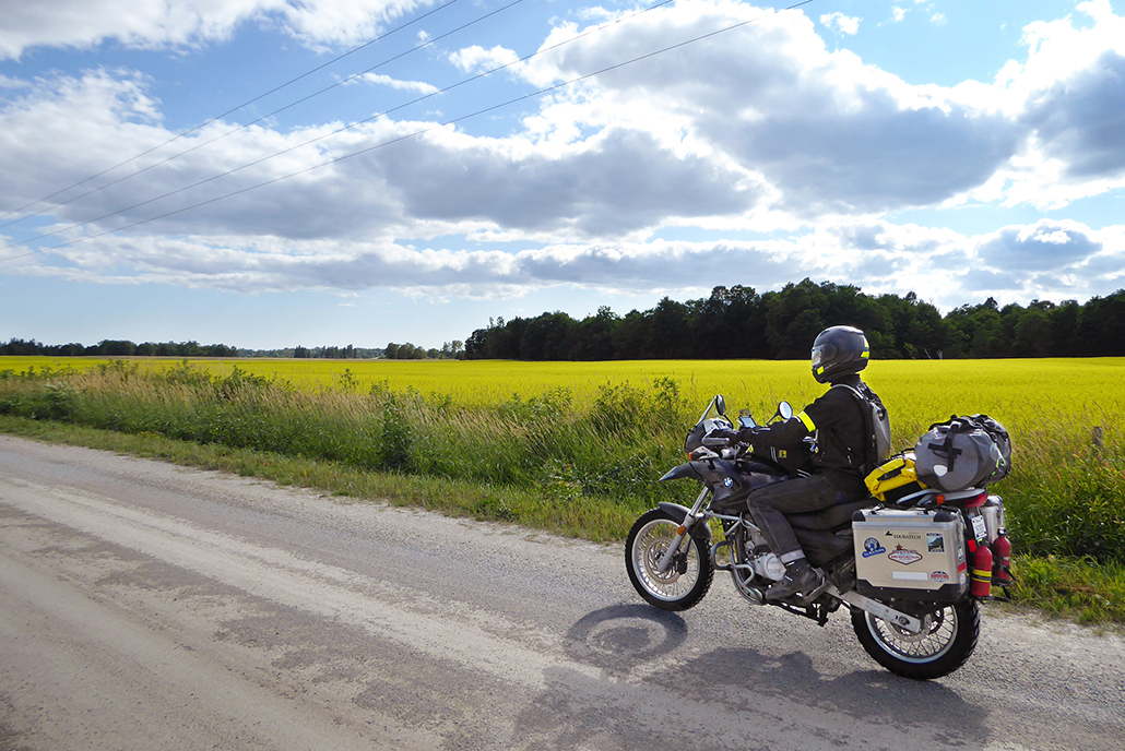 a motorcyclist riding a stretch of paved highway in front of a field of yellow canola blooming under a blue sky, with green summer forest on the horizon.