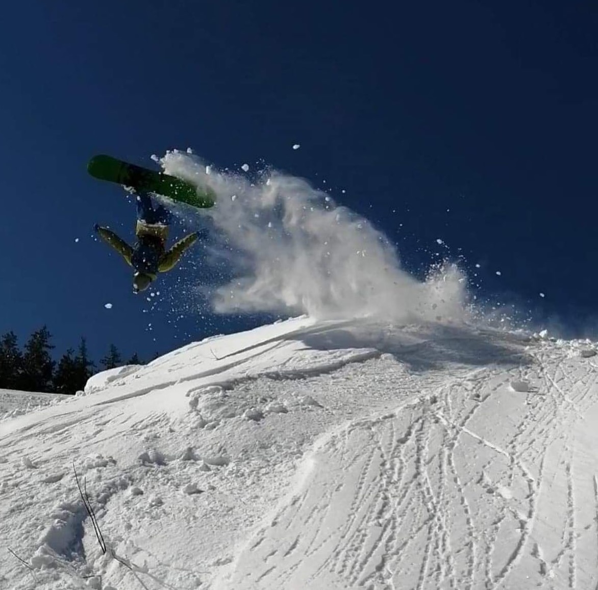 a snowboarder flips upside-down off a jump, followed by a blast of powder snow against a vivid blue sky.