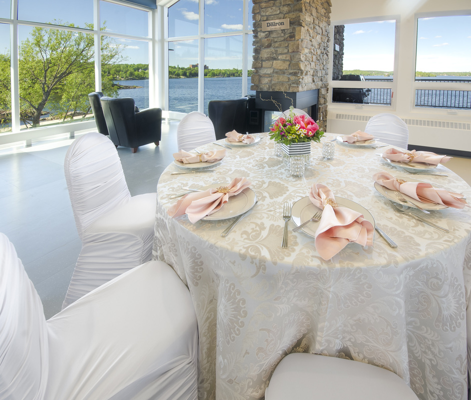 The Northern Water Sports Centre; an elegant table is set with white chairs and tablecloth, white plates, flowers and pink linen napkins. Shining floors meet a glass wall exposing a blue lake, sky and green trees outside.