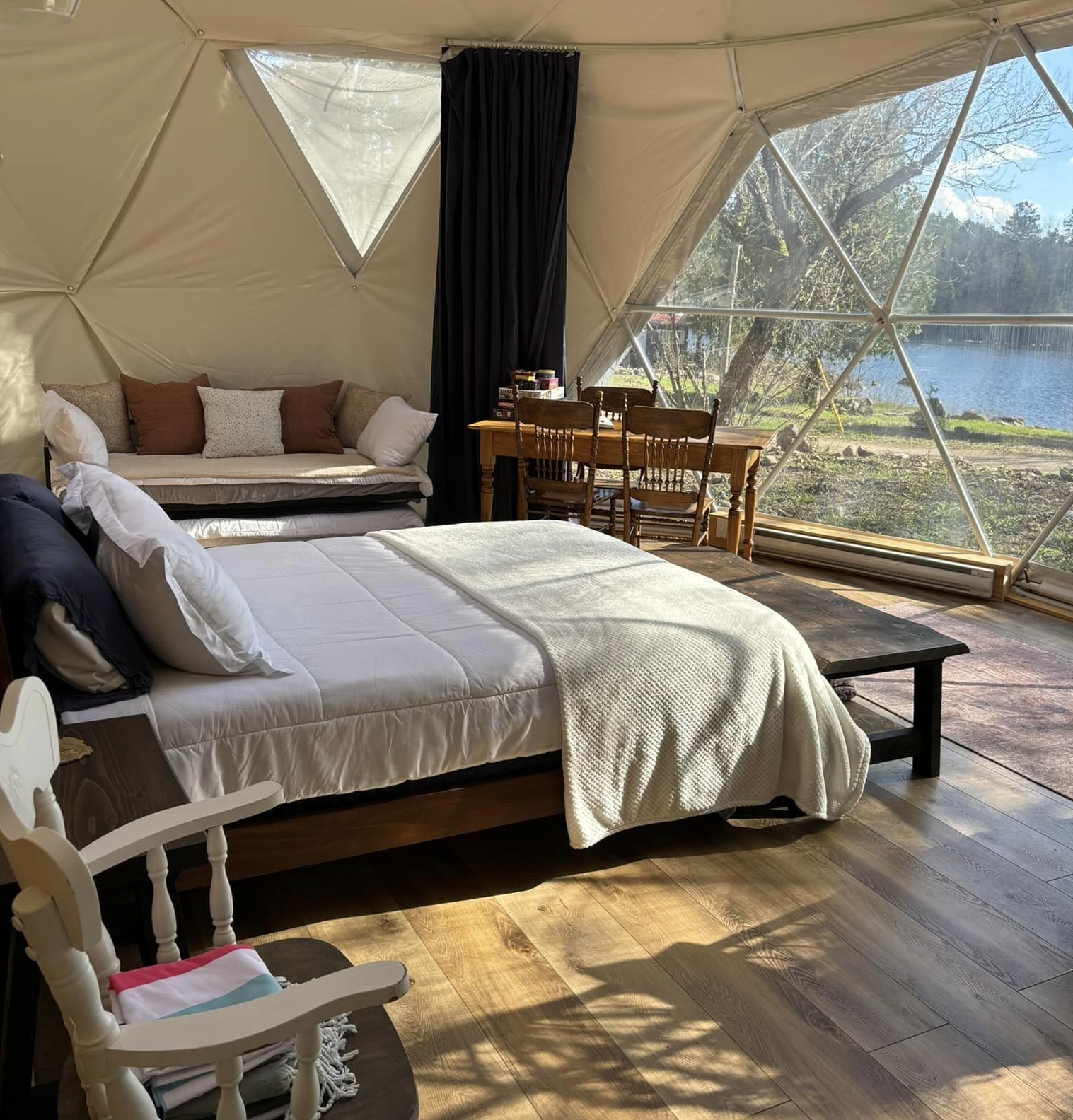 The interior of a sunny glamping dome with large windows, a double bed with comfortable blankets, a cusioned seat, and wooden table and chairs.