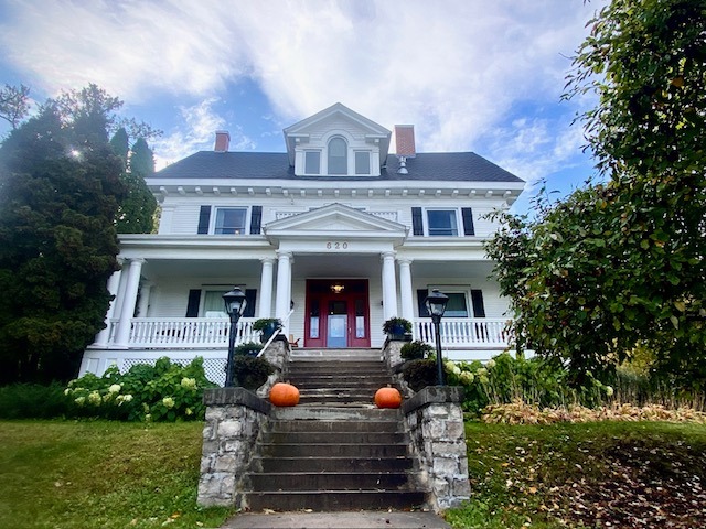 Presidents' Suites in the fall; a stately white two-storey mansion with stone steps up to the door, surrounded in large trees. Pumpkins sit on either side of the stairs.