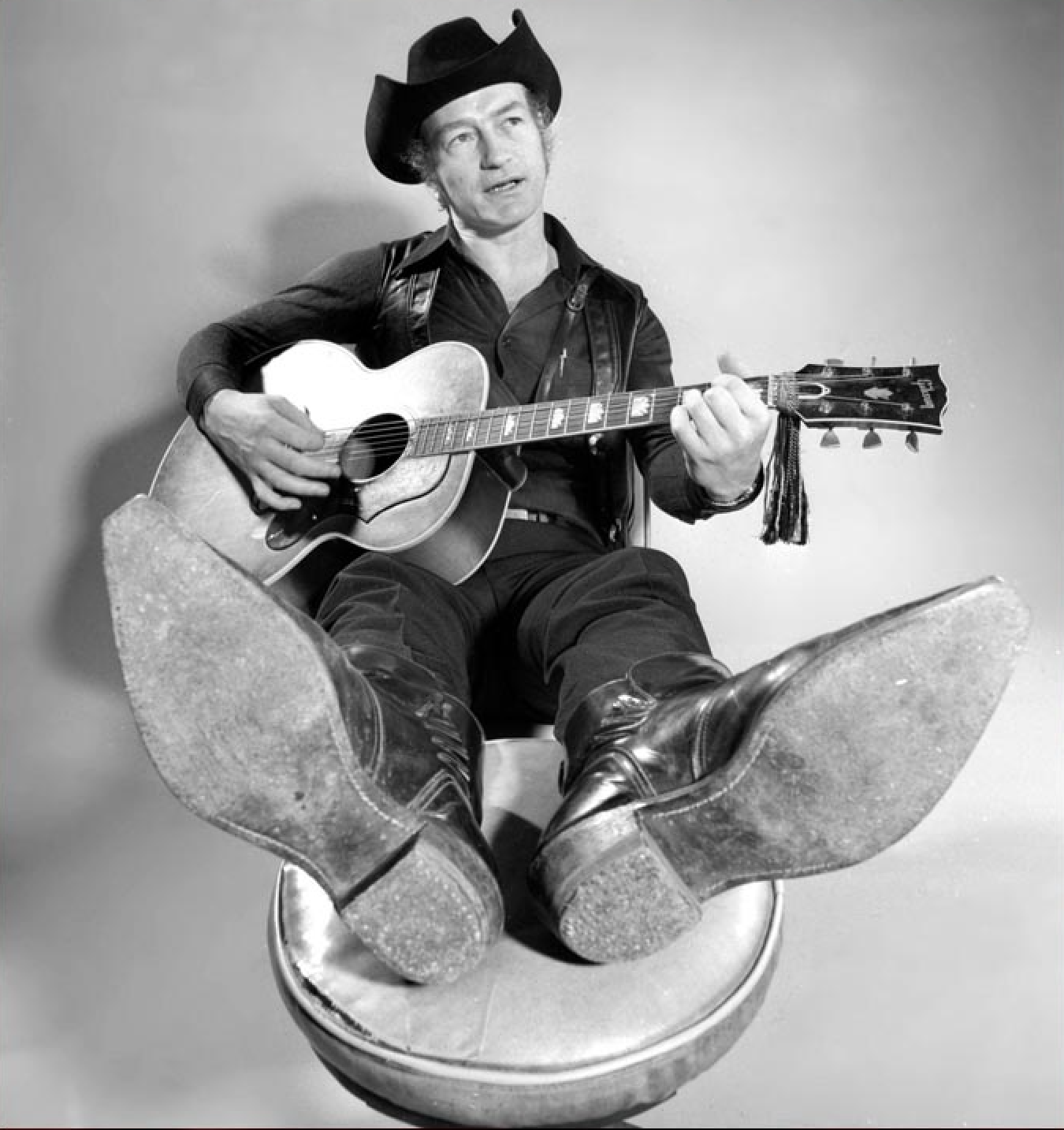 A black and white publicity photo of Stompin Tom Connors sitting on the floor playing his guitar.