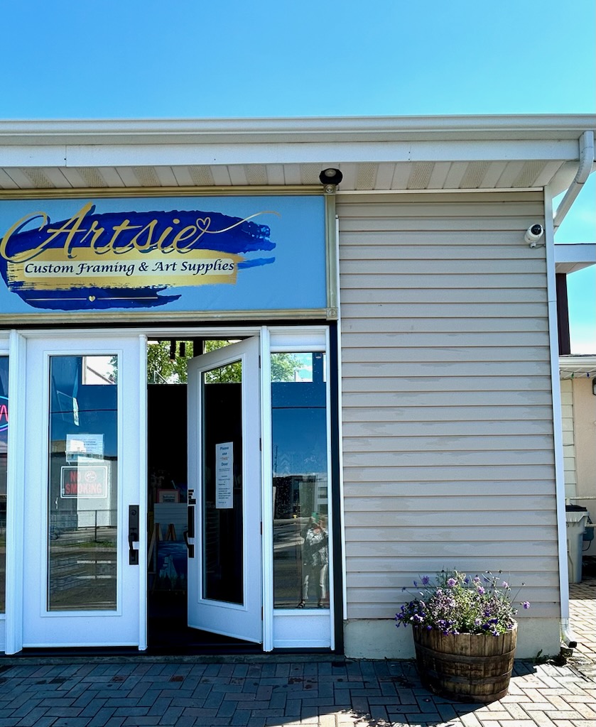 The exterior of the Artsie art supply store; a tidy beige building with white double doors and plants decorating the outside. 