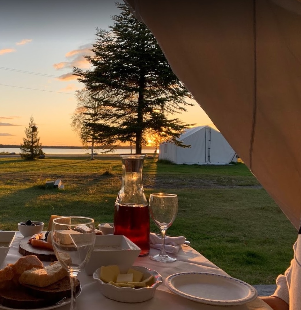 A meal at Wild Exodus; an outdoor table peeking out of a white canvas tent, spread with bread, wine and plates. Behind it is a green field and trees lit with a golden sunset.