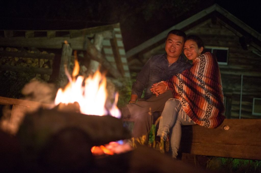 a couple cuddled together next to a campfire, smiling as they look into the flame.