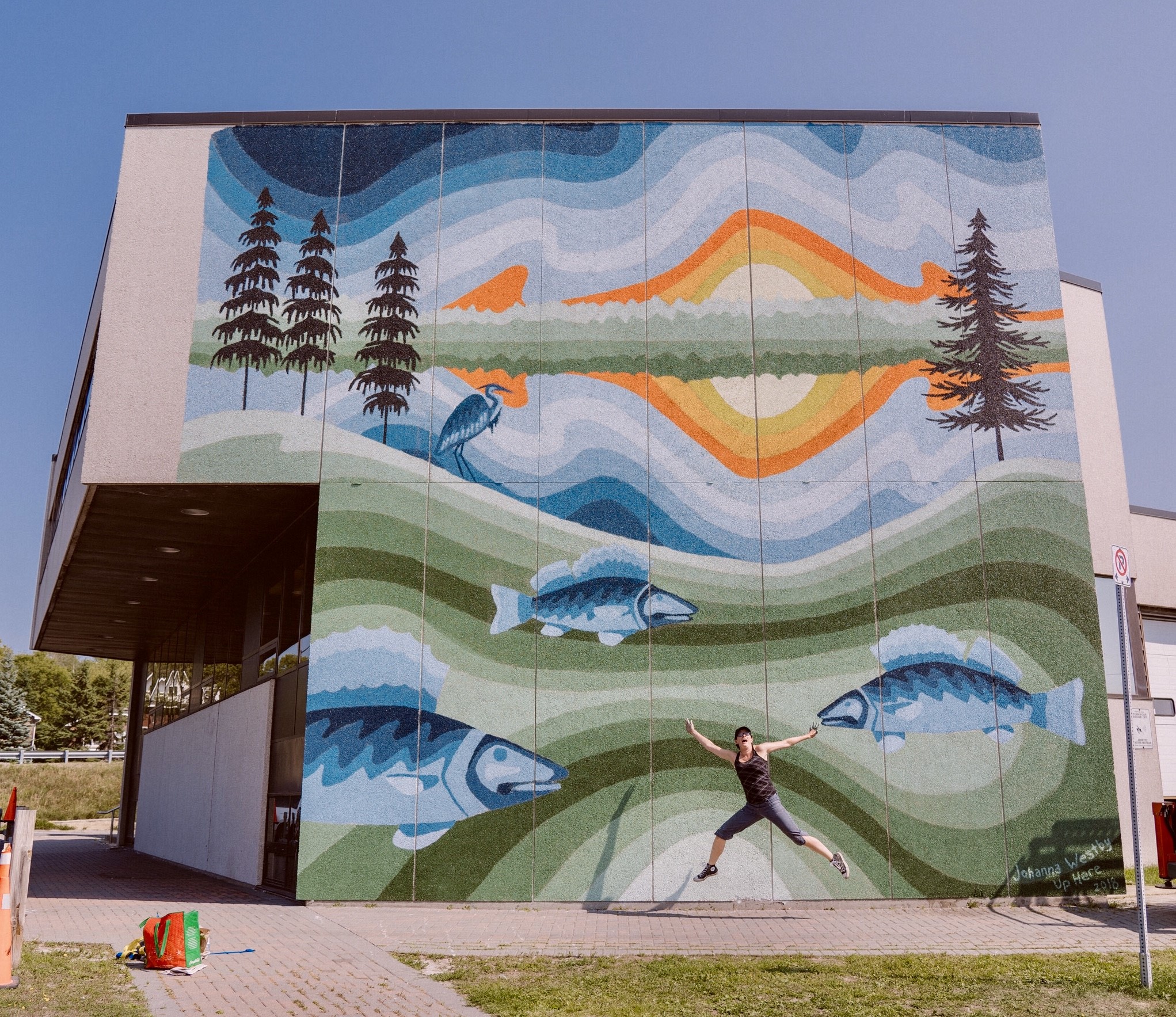 A colorful mural of nature on a brick wall in Sudbury Ontario. There is a woman in black jumping in front of it.