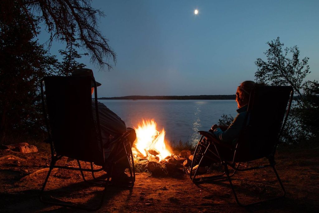 two people sitting next to a campfire beside a moonlit lake. 