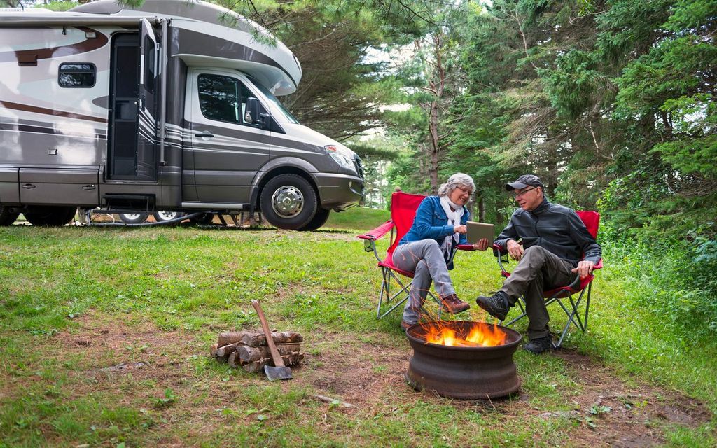 an older couple sitting next to a small campfire during the day, smiling together and looking at an iPad. 