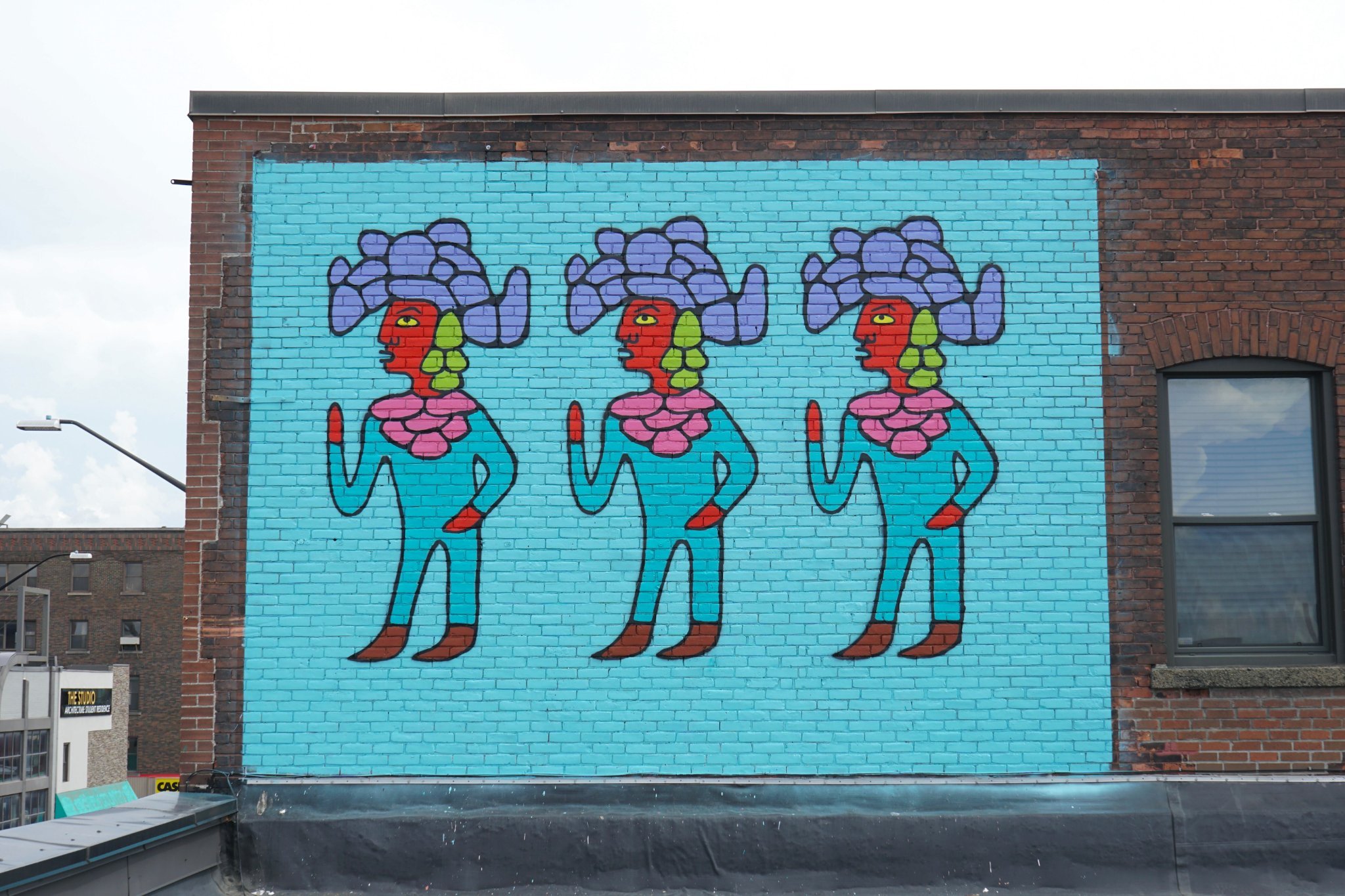 A turquoise, red and purple mural on a brick wall depicting 3 Indigenous figures in the Woodland artist style.