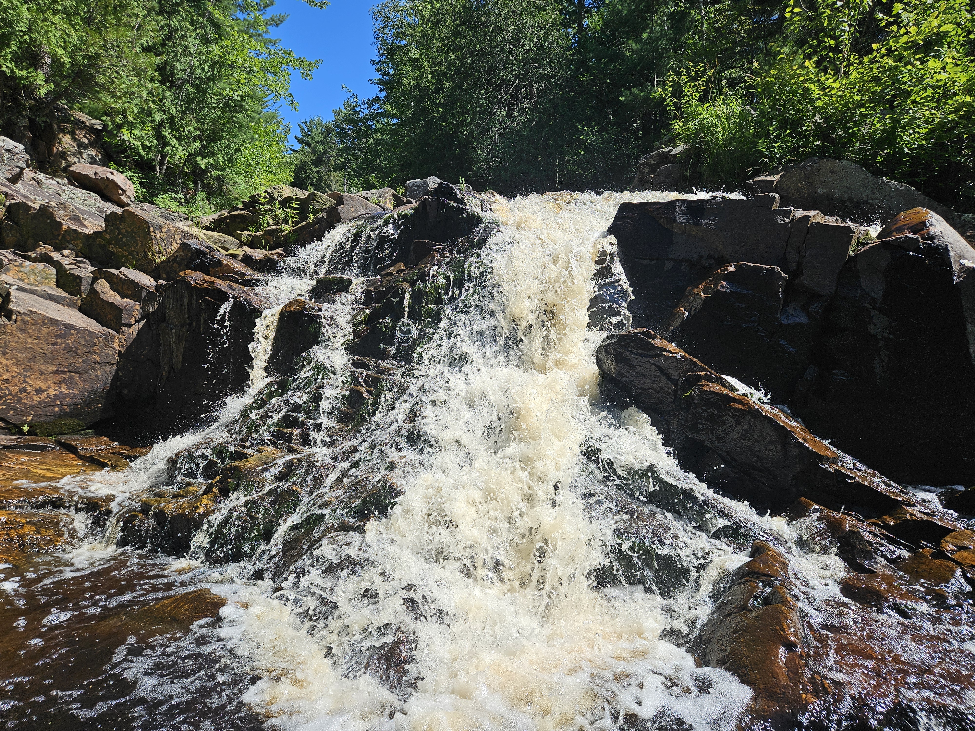 Duchesnay Falls; a cascading waterfall down a multi-levelled rock cliff surrounded by lush green trees under a bright blue sky.