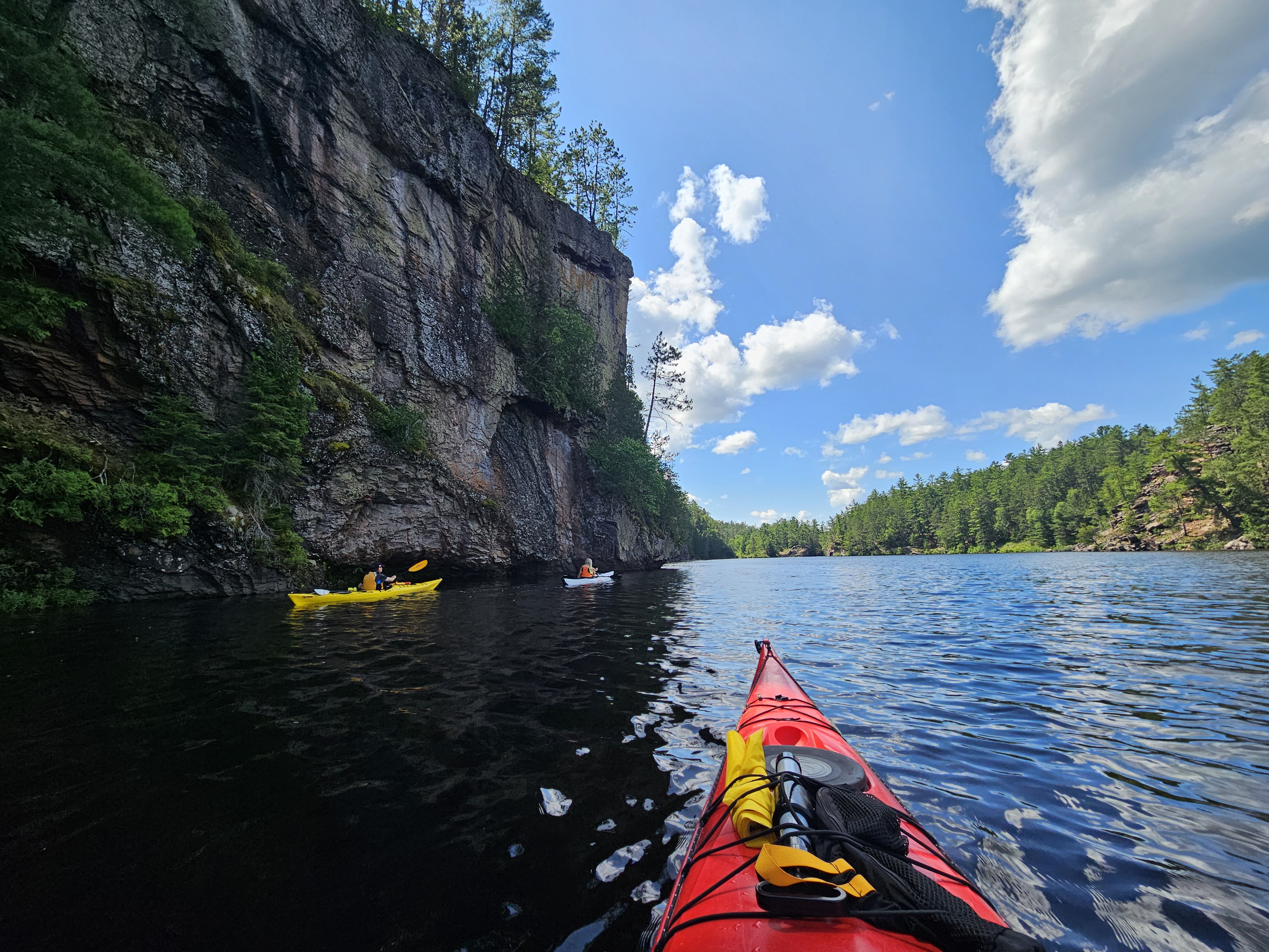 3 kayakers paddle along a tall, forested rock cliff on the Mattawa River on a sunny summer day.