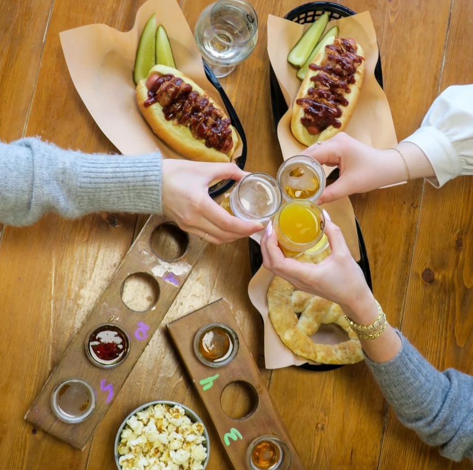 3 people clink their drinks together in the centre of a wooden table, over top of popcorn, pretzels and chili dogs.