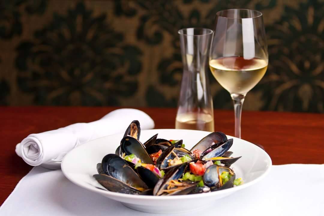 A plate of mussels on a red table cloth next to a decanter of white wine with and a glass.