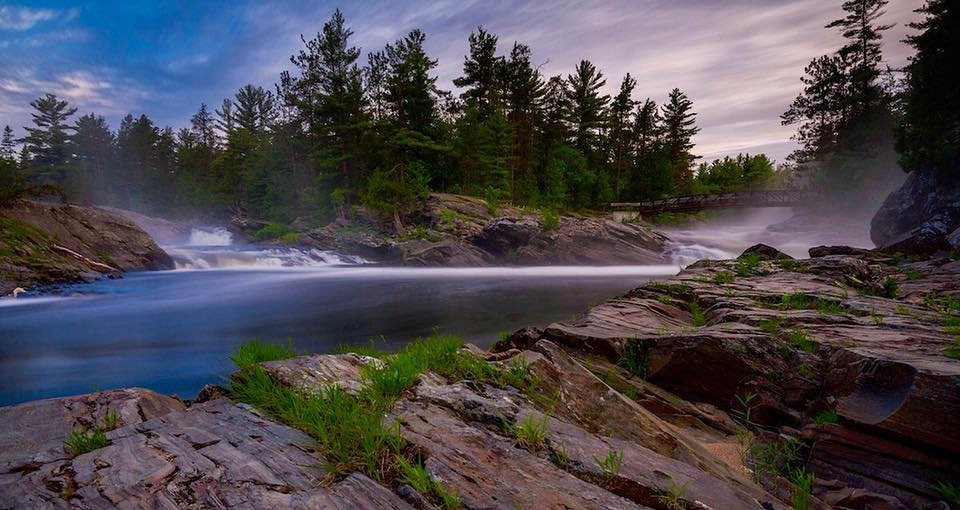 the glassy Aux Sables River at the base of Chutes falls, reflecting a dusky sky. The river has smooth rock banks and is surrounded by pine forest. 