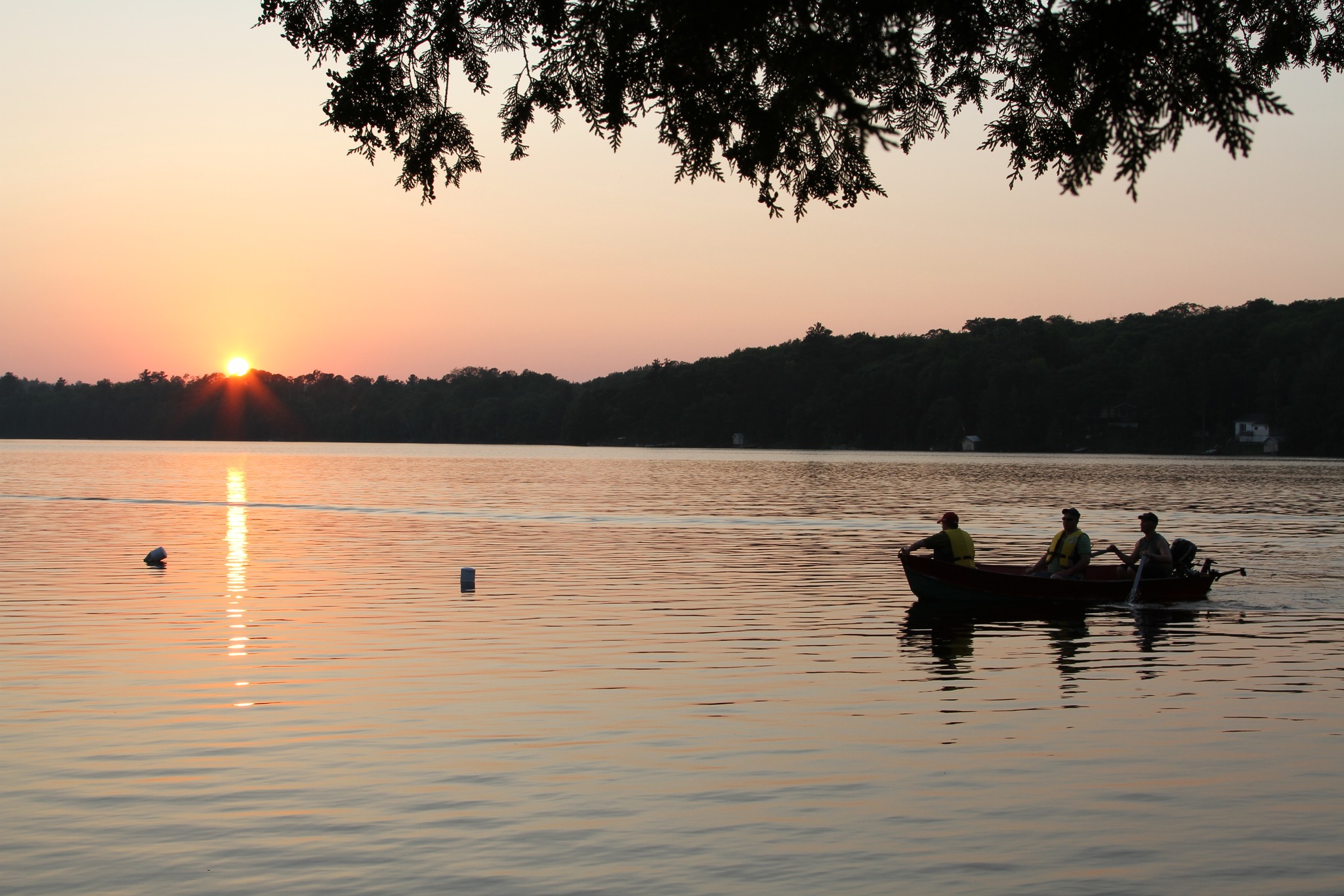 a boat holding 3 people who are fishing, floating on a glassy lake at sunset. A band of forest is silhouetted on the horizon.