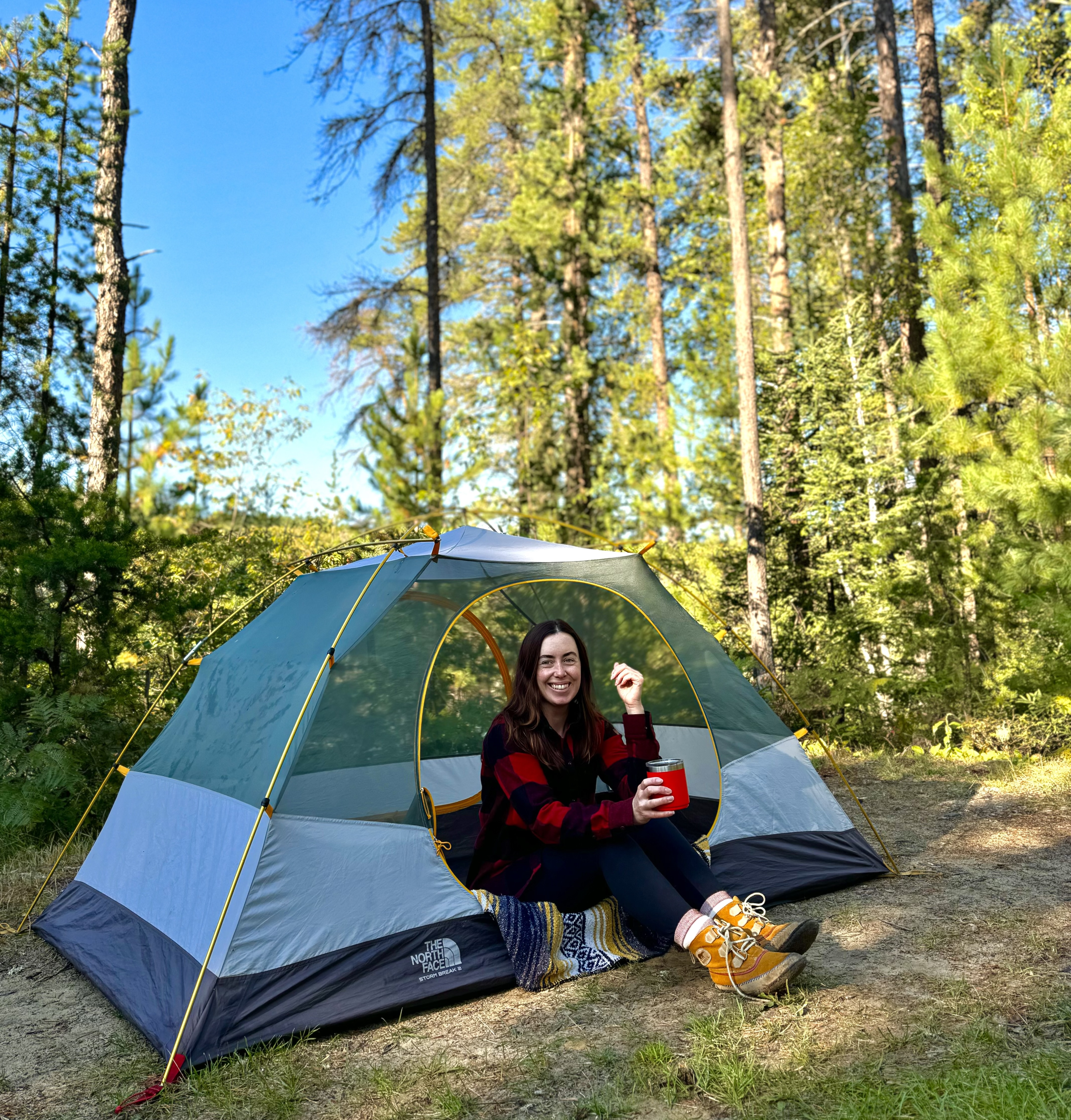 a smiling woman wearing hiking boots, sitting in the doorway of a small tent, in front of a tall green pine forest and brilliant blue sky.