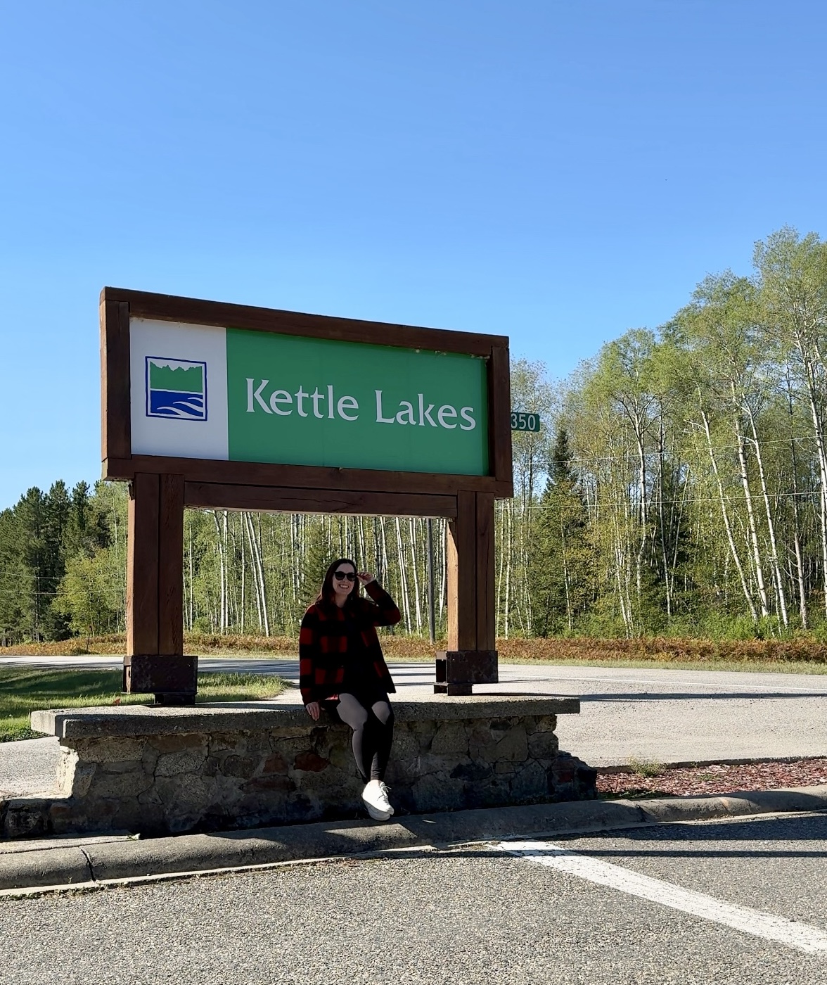 A woman sitting at the base of a large Ontario provincial parks sign that says "Kettle Lakes" at the side of a paved road. The sky is bright blue and there is green forest in the background. 