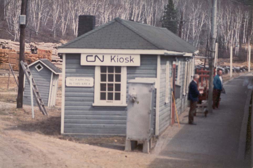 Kiosk Station; a small wooden train station next to a train track, surrounded by forest. Two people stand on the wooden platform in front of it. 