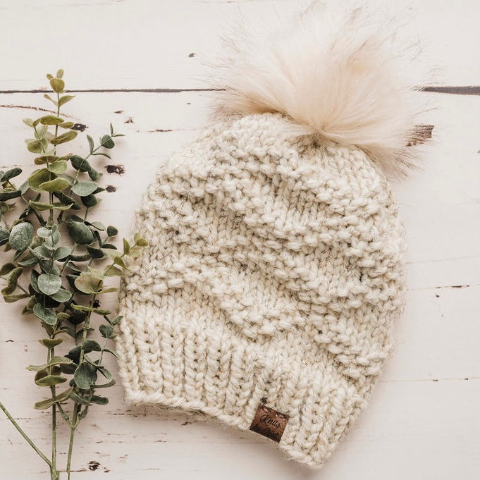 a white handkitted toque with diamond pattern and a fur pom pom.