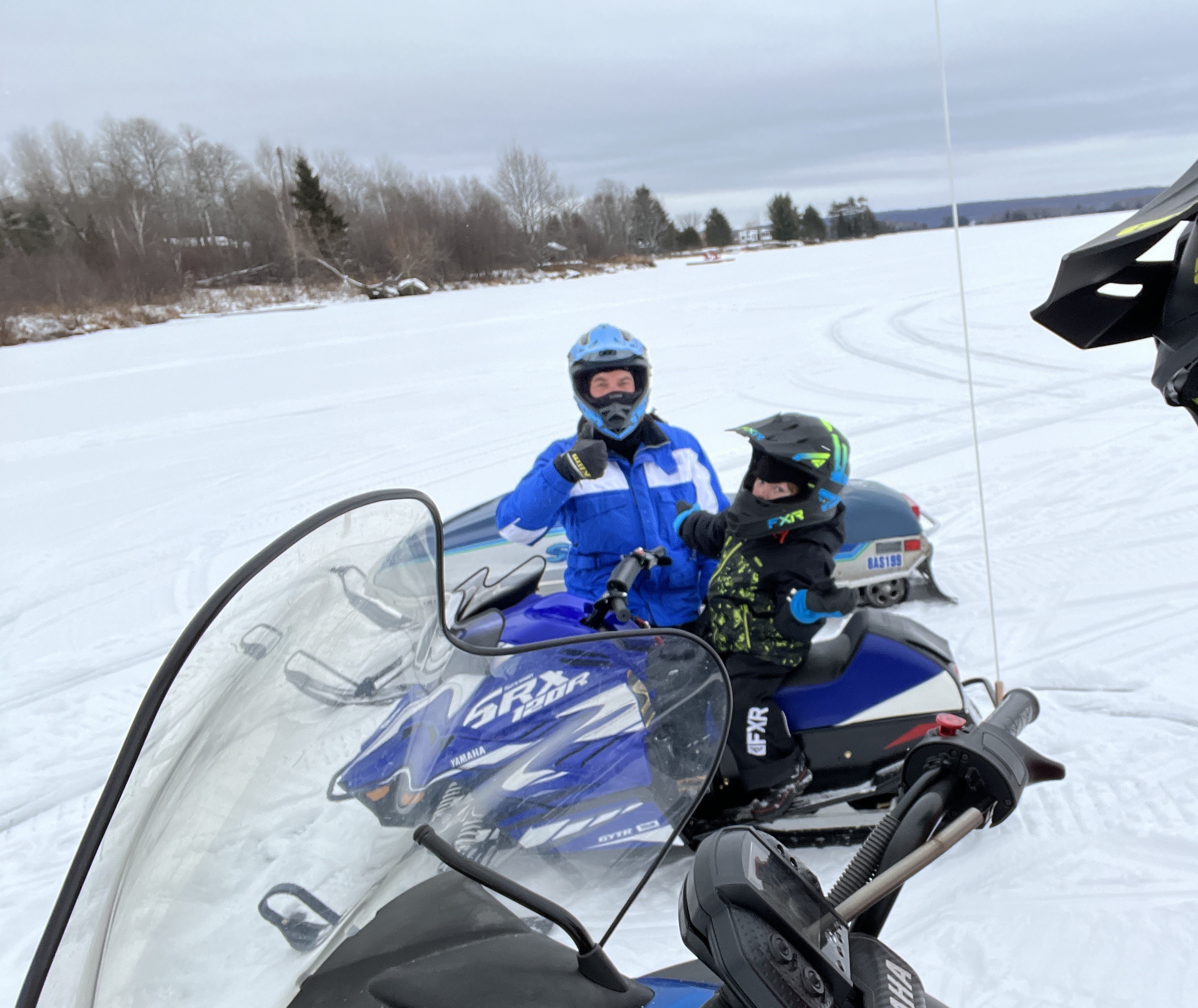 A man and small boy on their snowmobiles in a snowy field on a cloudy day.