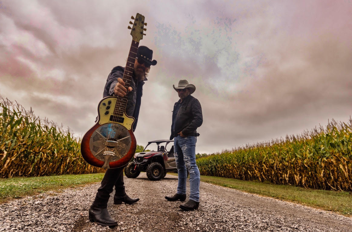 two men in cowboy hats and sunglasses stand on a gravel road in a corn field, in front of an ATV. One man holds up a guitar. 