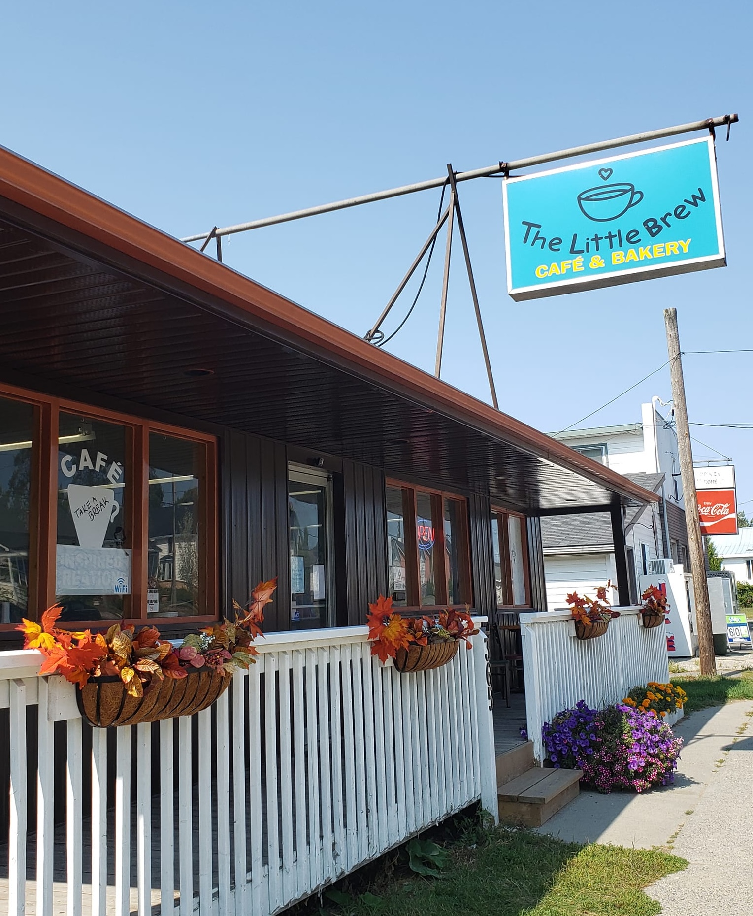 Little Brew Café; a pretty cafe with windows facing the street and a white wood-rail patio in front. A turquoise "Little Brew Café" sign hangs overhead, featuring a teacup with a heart above it.