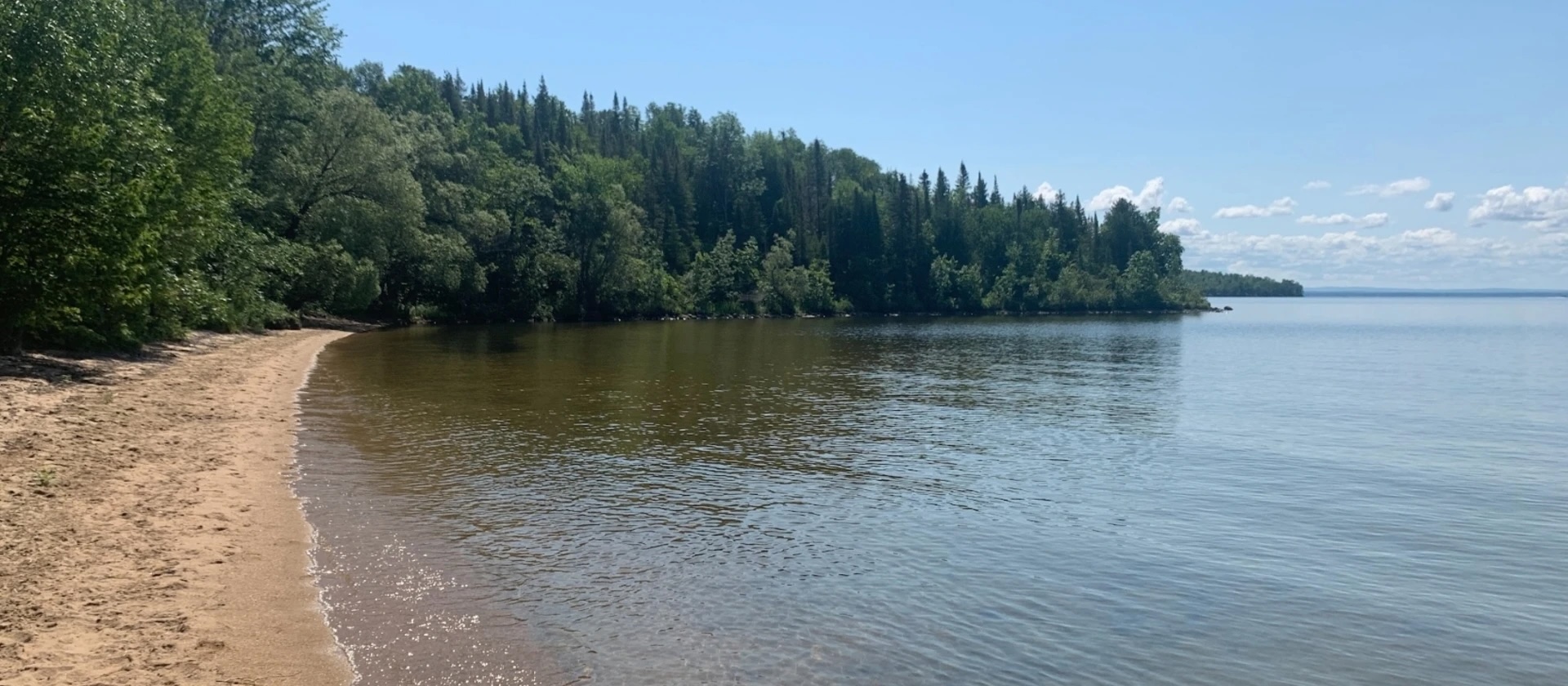 a long strip of sandy beach along a glassy lake, skirted with dense green forest.