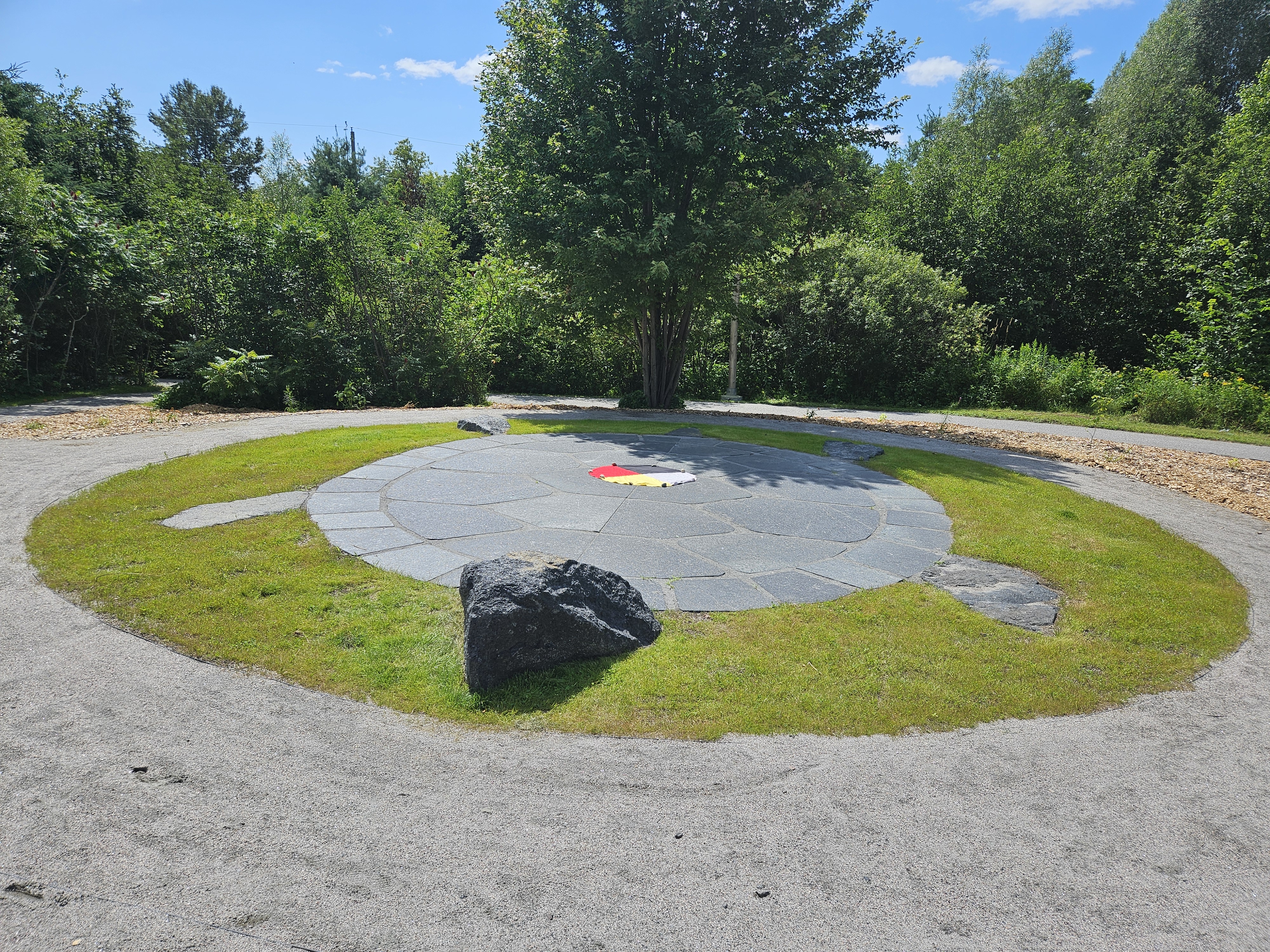 Miskwaadesi; a large stone turtle with a medicine wheel in the centre, set into ac circle of green grass, surrounded by a stone walkway and green trees. 