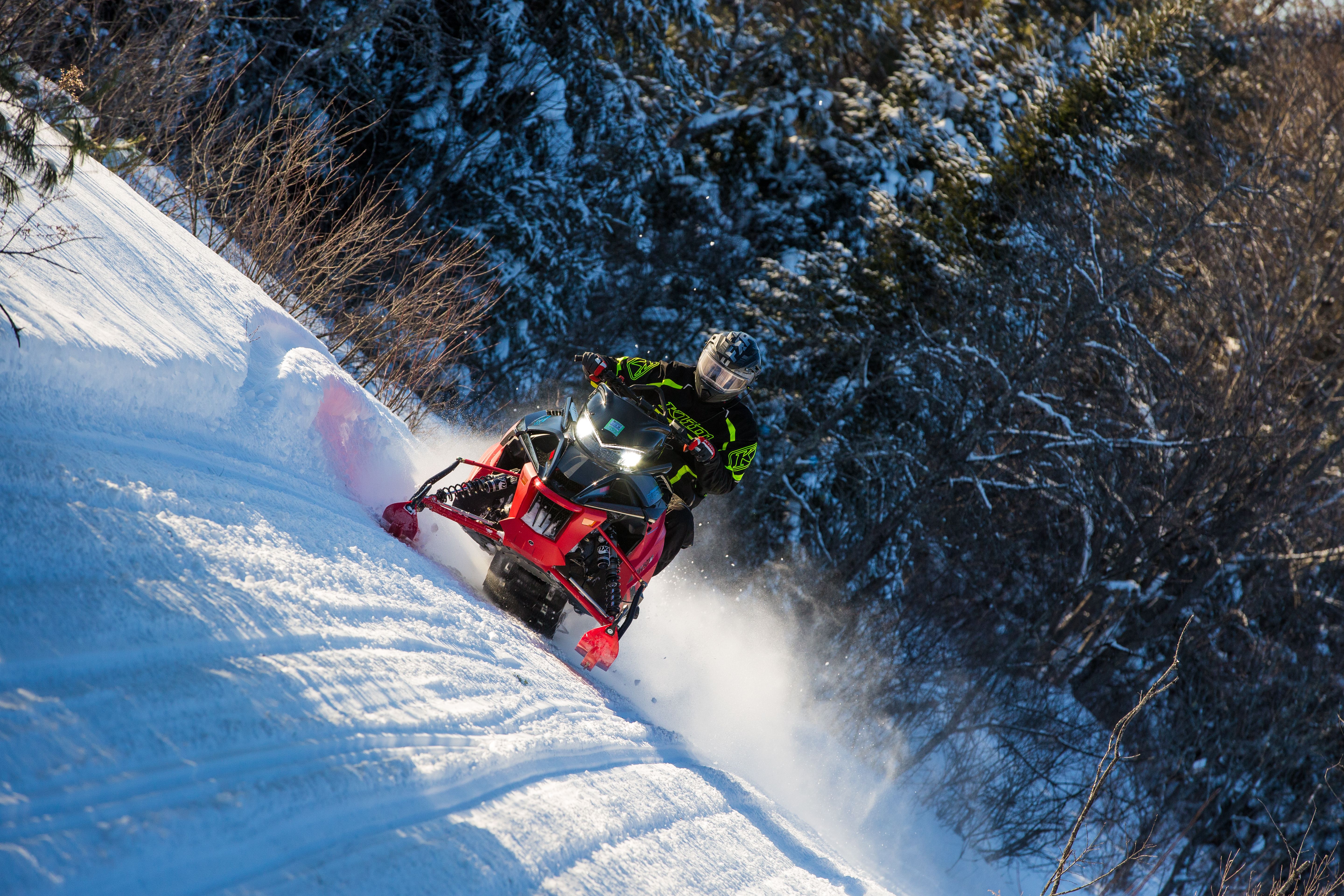 a snowmobiler rides down a powdery trail surrounded by snowy spruce forest on a sunny day.