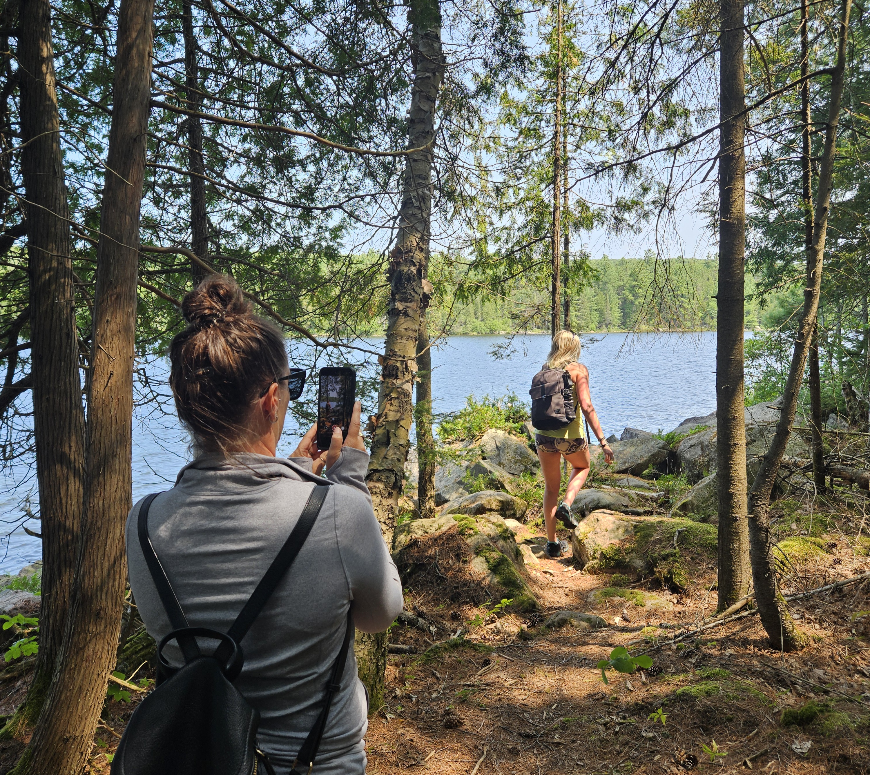 a woman snaps a photo as her friend walks along a series of large boulders along a lakeside under a canopy of tall pine trees.