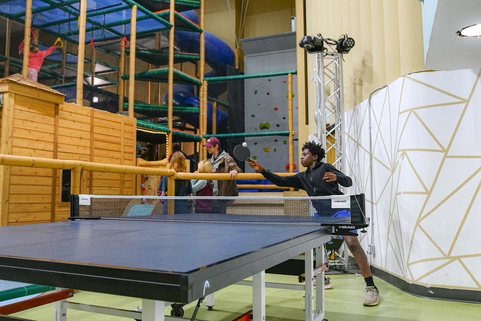 kids of all ages playing in a large indoor play park, with high play equipment that goes to the ceiling. One in the foreground is playing ping pong. 