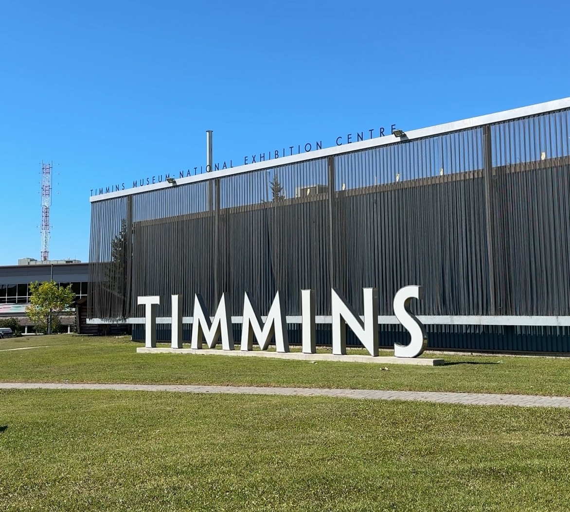 A sign of free-standing metal letters that spells "TIMMINS", set on green grass under a bright blue sky, outside of the Timmins Museum Nation Exhibition Centre. 