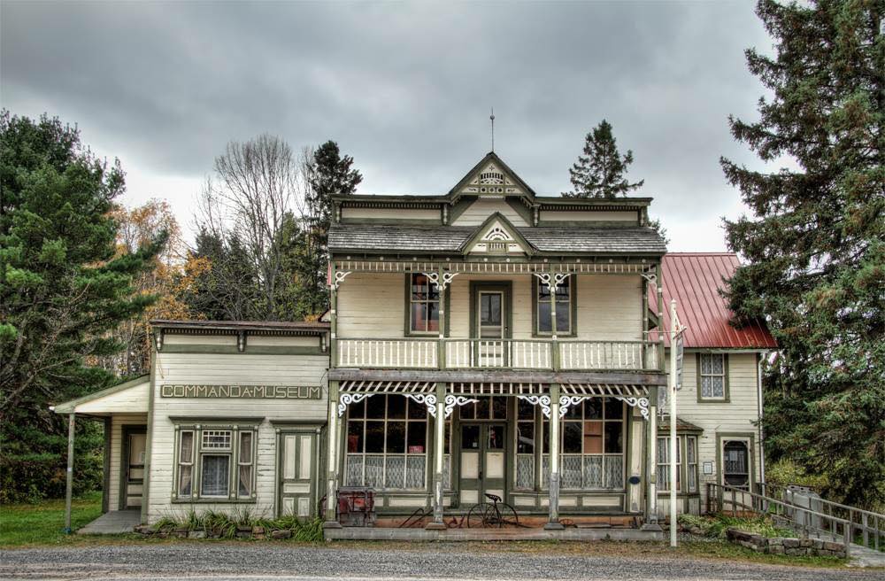 The Commanda Museum; an antique, bi-level grocery store with white siding and covered wooden porch. Dark clouds loom overhead giving the store a sense of foreboding. 