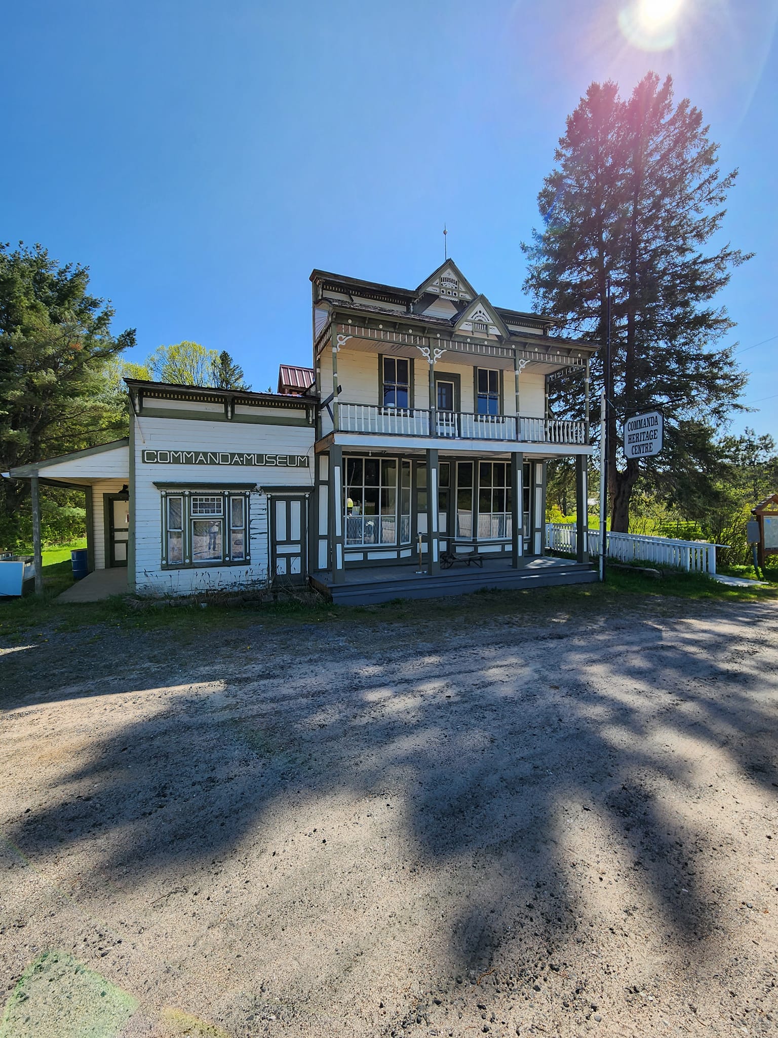 The Commanda Museum; a white antique bi-level grocery store with a covered porch, the sun behind it casting a long jagged shadow in front. 