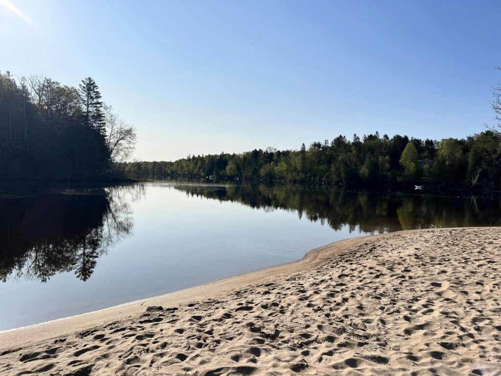 a white sandy beach at the mouth of the Aux Sables river, skirted by dense forest under a clear blue sky.