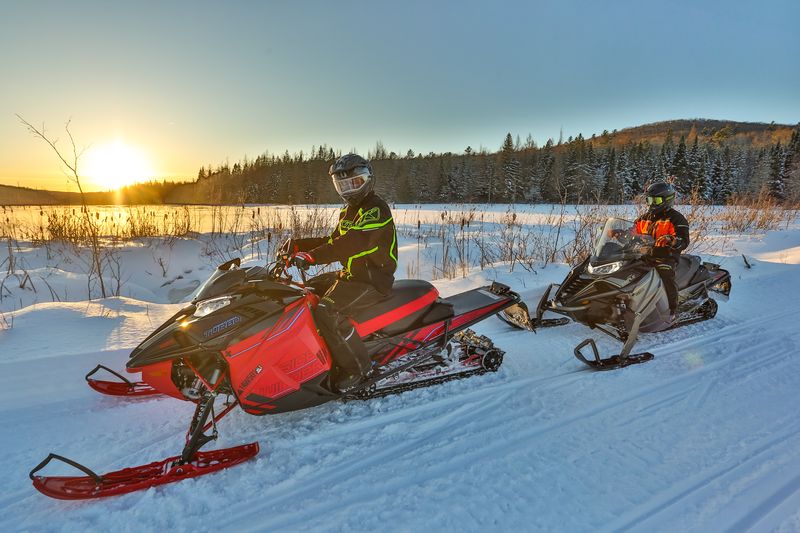 Two riders on their snowmobiles parked on a snowy trail looking at a sunrise on a clear morning.