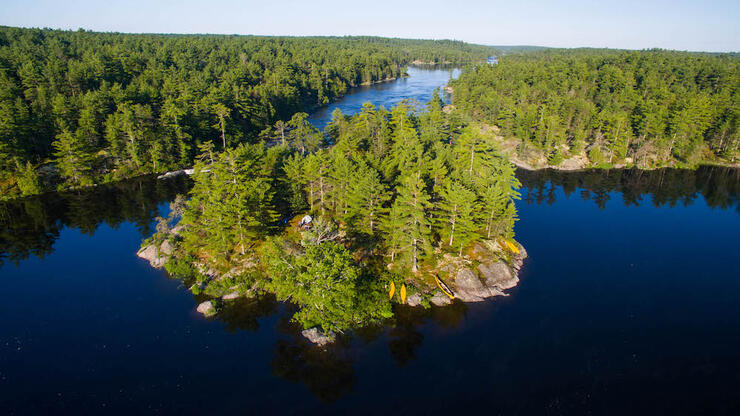 a wide, calm blue lake fed by a river the winds into the distance, through thick green boreal forest. In the middle of the lake is a rocky, tree-covered island with canoes and a tent on it.