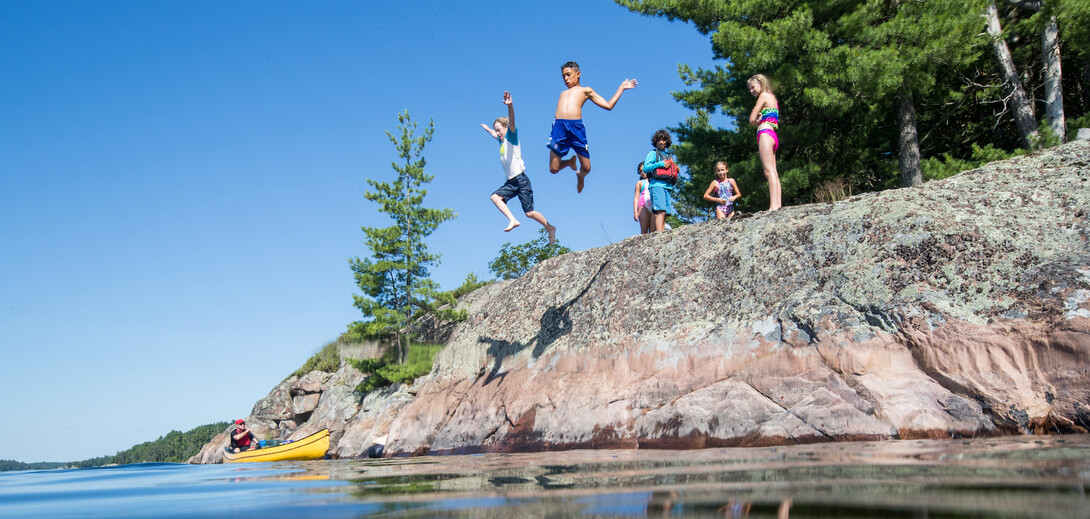 Children in bathing suits jumping happily off a tall, forested rock bank into the French River on a clear summer day.