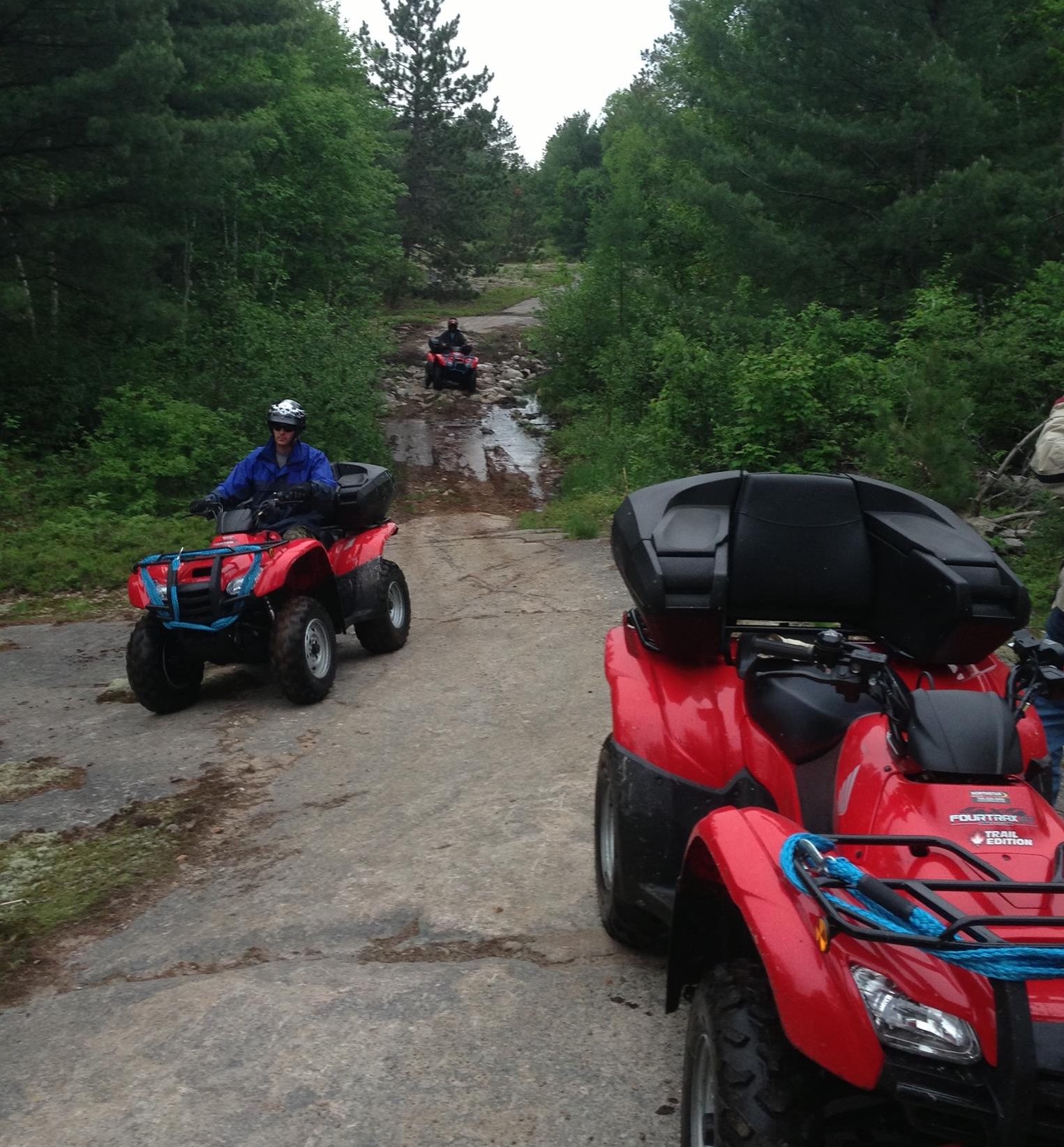 A line of riders on ATVs drive up a rocky forest trail on a rainy day.