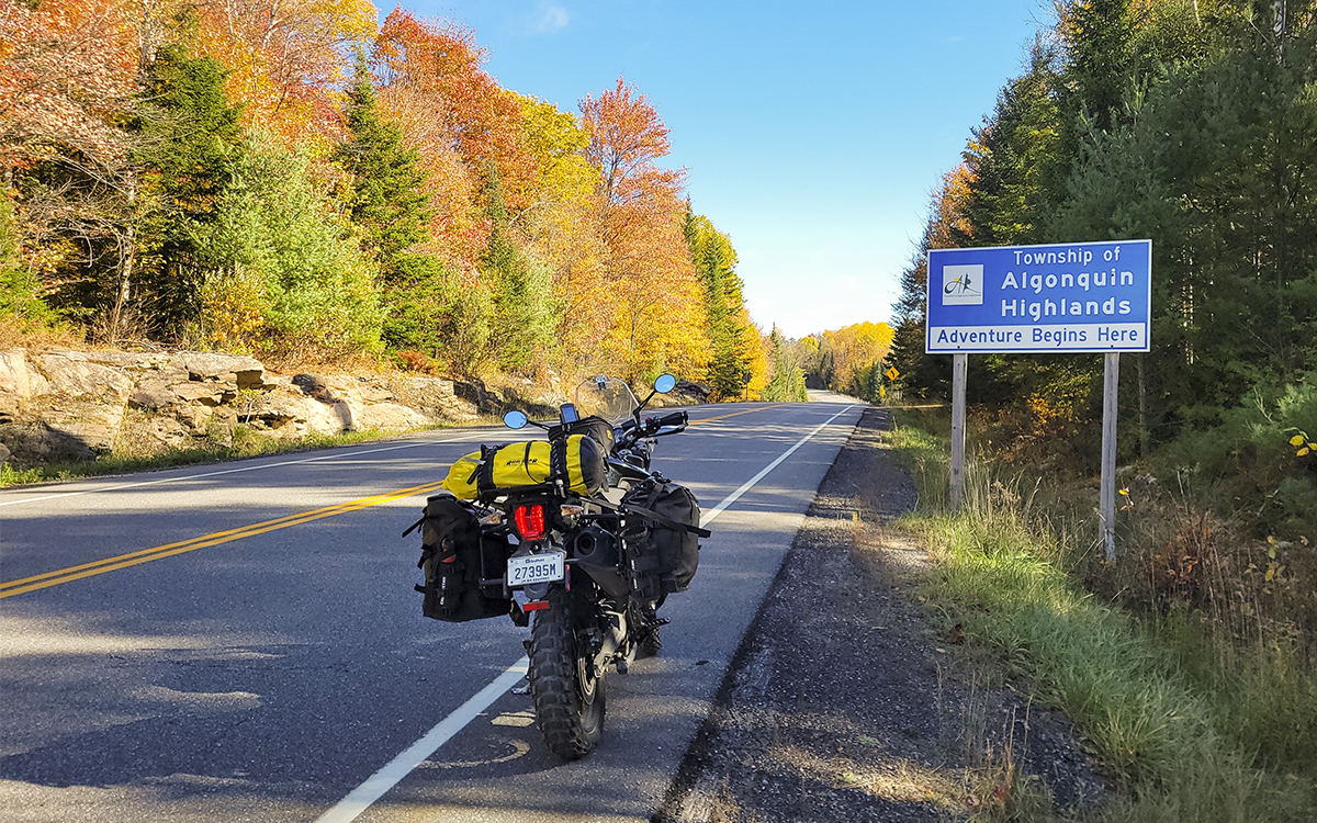 a motorcycle parked at the edge of a paved road skirted by colourful autumn forest. A blue sign by the side of the road reads "Township of Algonquin Highlands—Adventure Begins Here". 