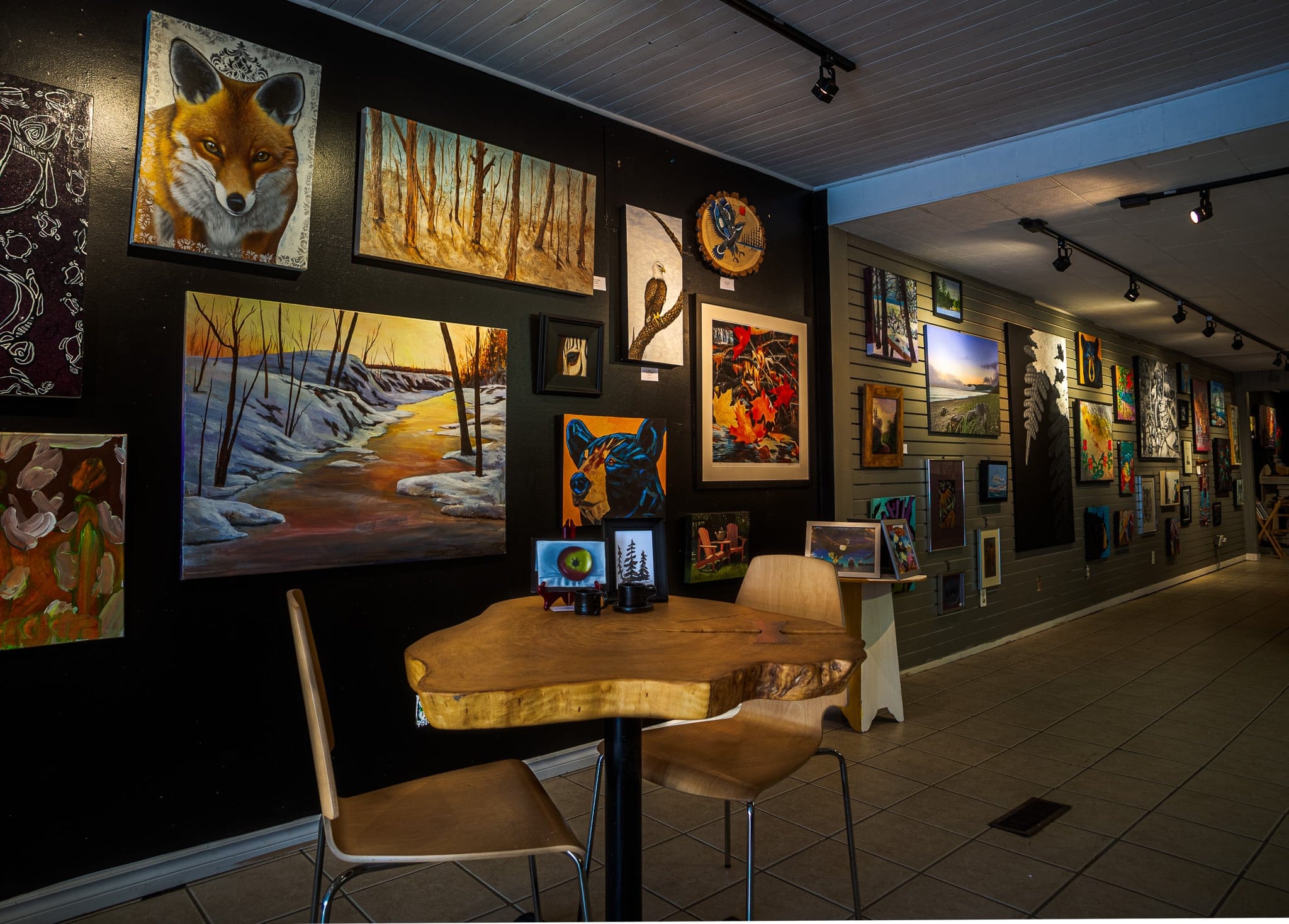 A wood cafe table and chair in front of a long gallery wall of beautiful paintings at Backstreet Gallery.