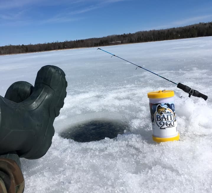 the feet of a boot-wearing ice fisher louging next to a hole in the ice on a sunny day, an ice fishing rod and Bait2Go bucket next to the hole.