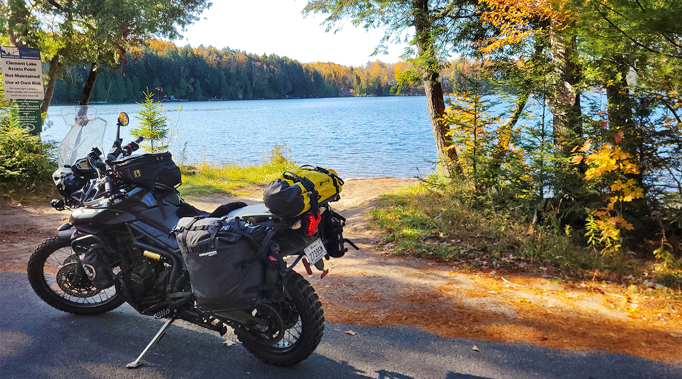 a motorcycle parked at the edge of a road next to a calm blue lake surrounded by autumn forest on a sunny day.