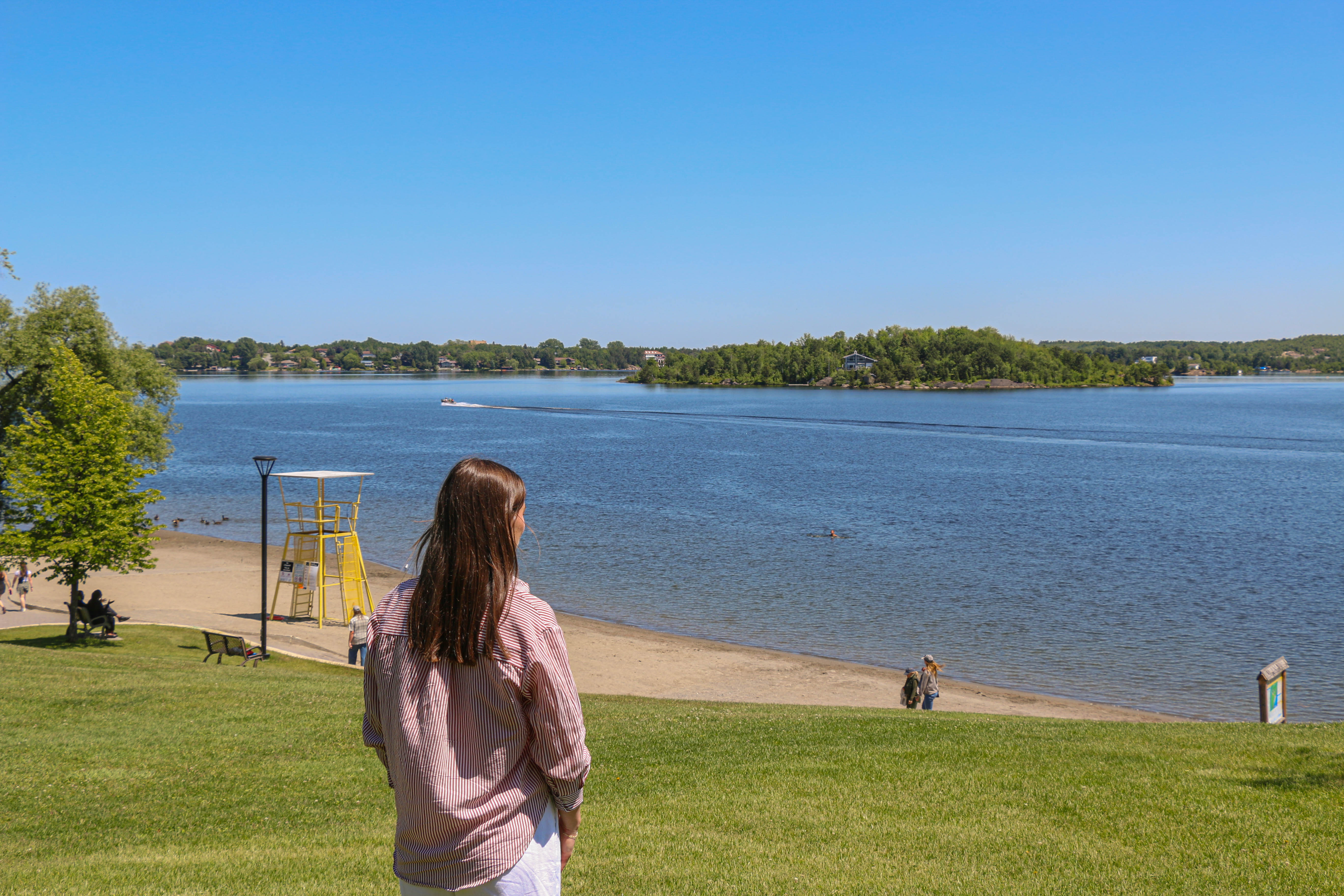 Bell Park Beach; a girl stands on a green grassy hill on a summer day, looking out over a calm blue lake, edged in sandy beach under a clear blue sky. A lifeguard chair sits at one end of the beach, and there are people sitting on a park bench near the water. 