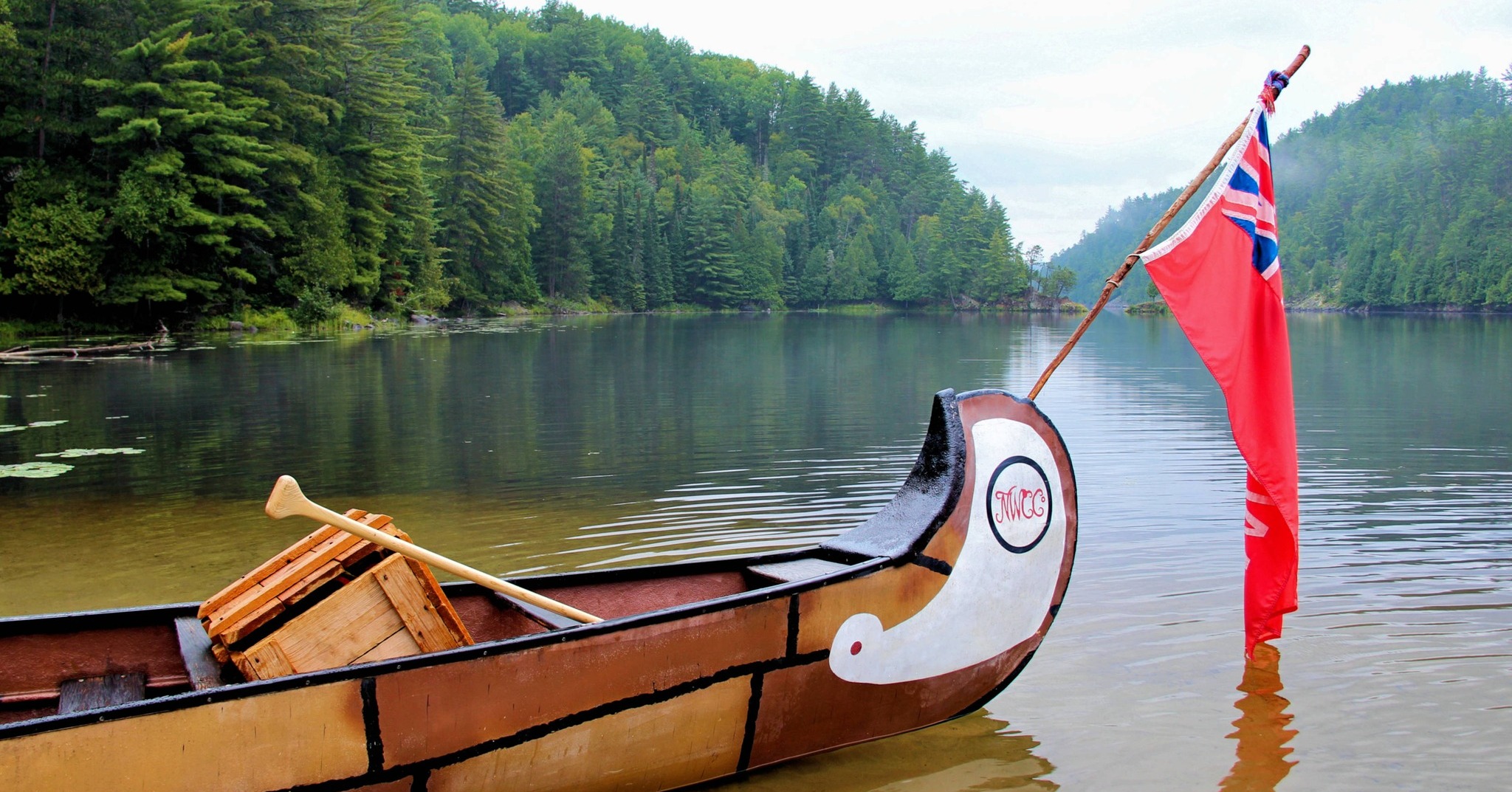 a large wooden reenactment canoe with a wooden crate and paddle in it, floating in the glassy French River, surrounded by dense green forest. The canoe is flying the Red Ensign flag of early Canada.